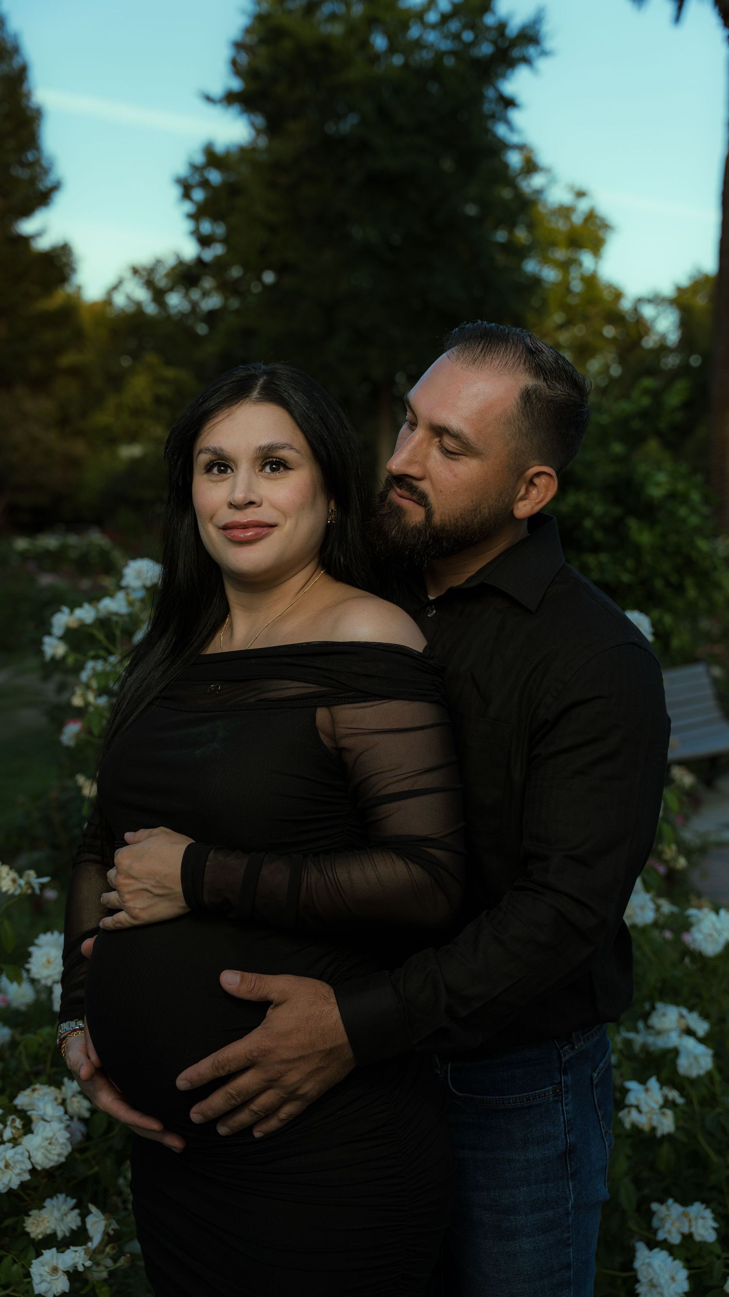 Intimate maternity photo in black dress against evening garden setting.
