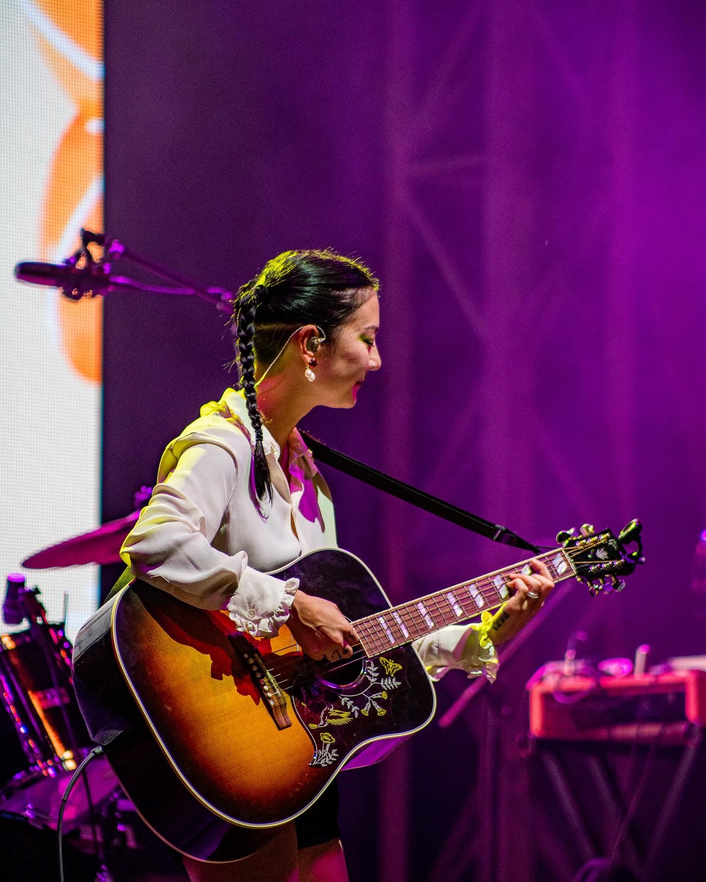 Guitarist performing on stage with sunburst acoustic guitar against purple backdrop.