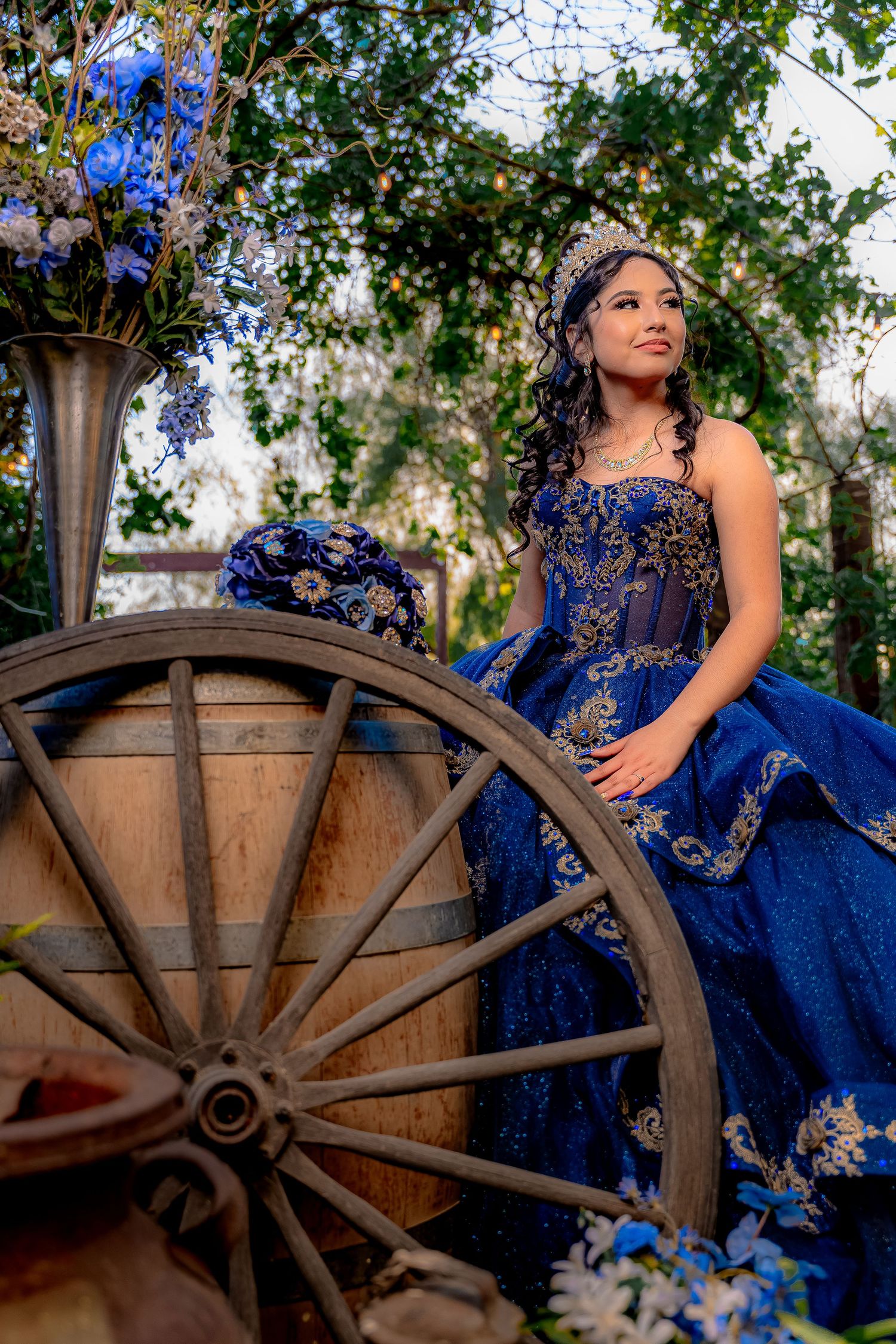 Elegant outdoor portrait with vintage wagon wheel and blue gown against natural foliage.