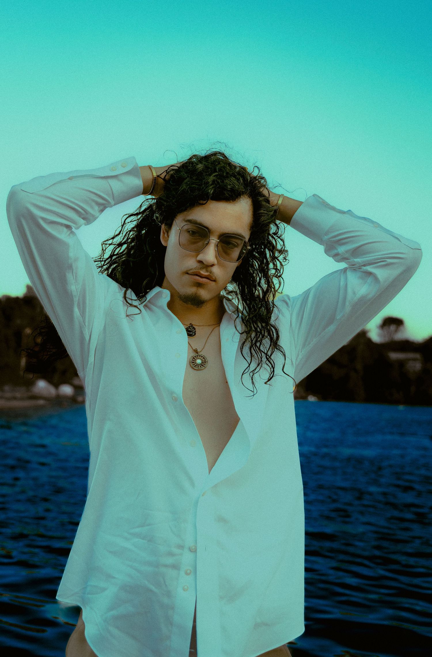 Figure in white button-down shirt with long curly hair and sunglasses poses by waterfront at sunset.