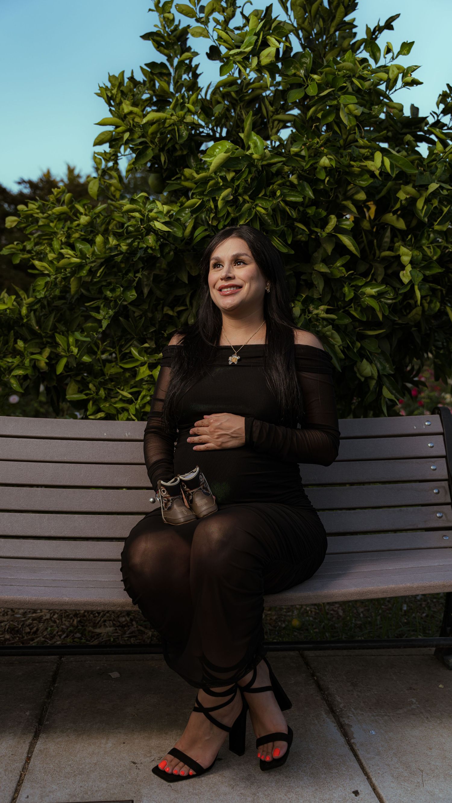 A figure in black clothing sits on a wooden bench surrounded by lush green foliage in an outdoor setting.
