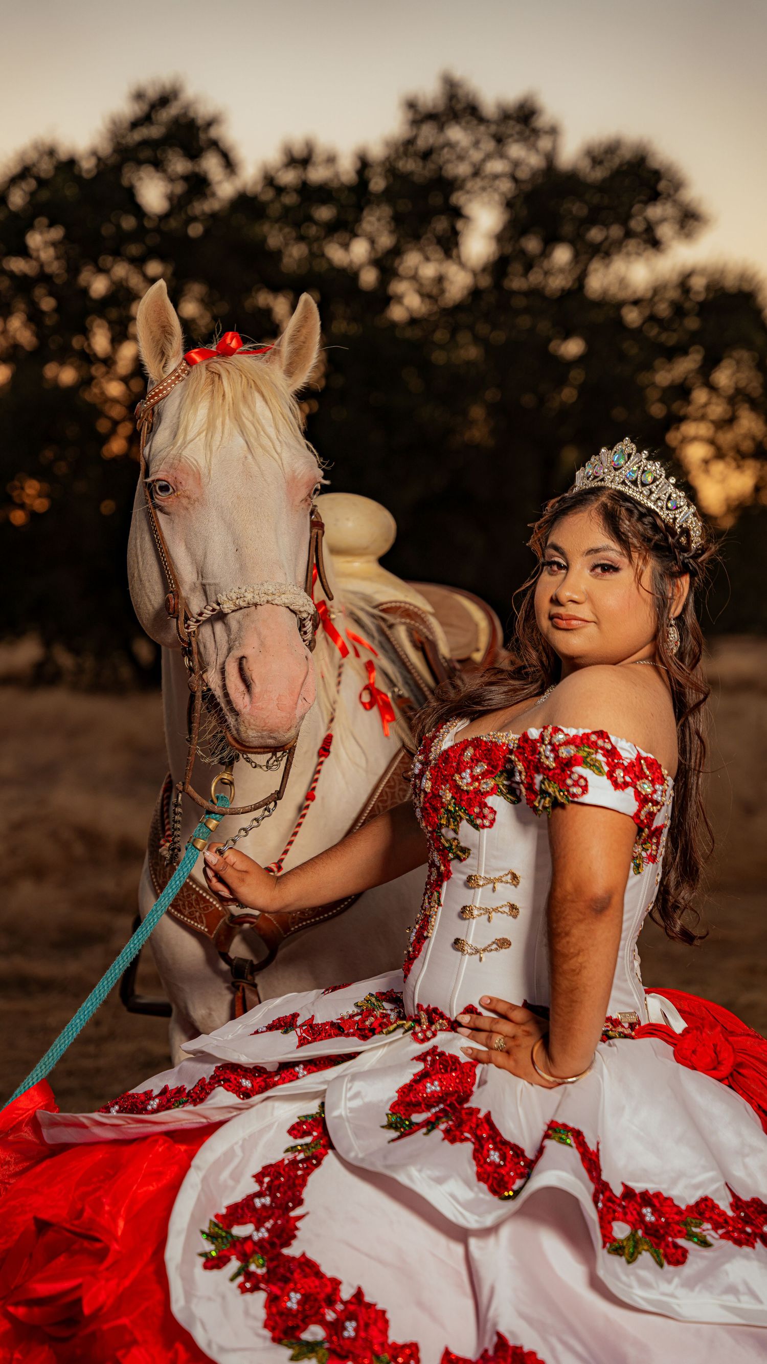 A white and red quinceañera gown sparkles as its wearer poses with a white horse at dusk.