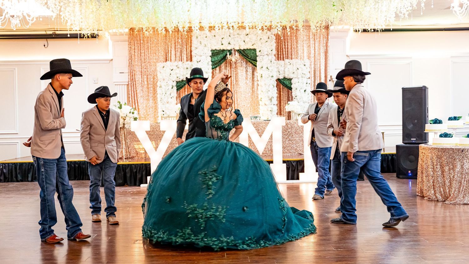 Dancers in cowboy hats perform at a quinceañera celebration in a decorated ballroom with pink lighting.