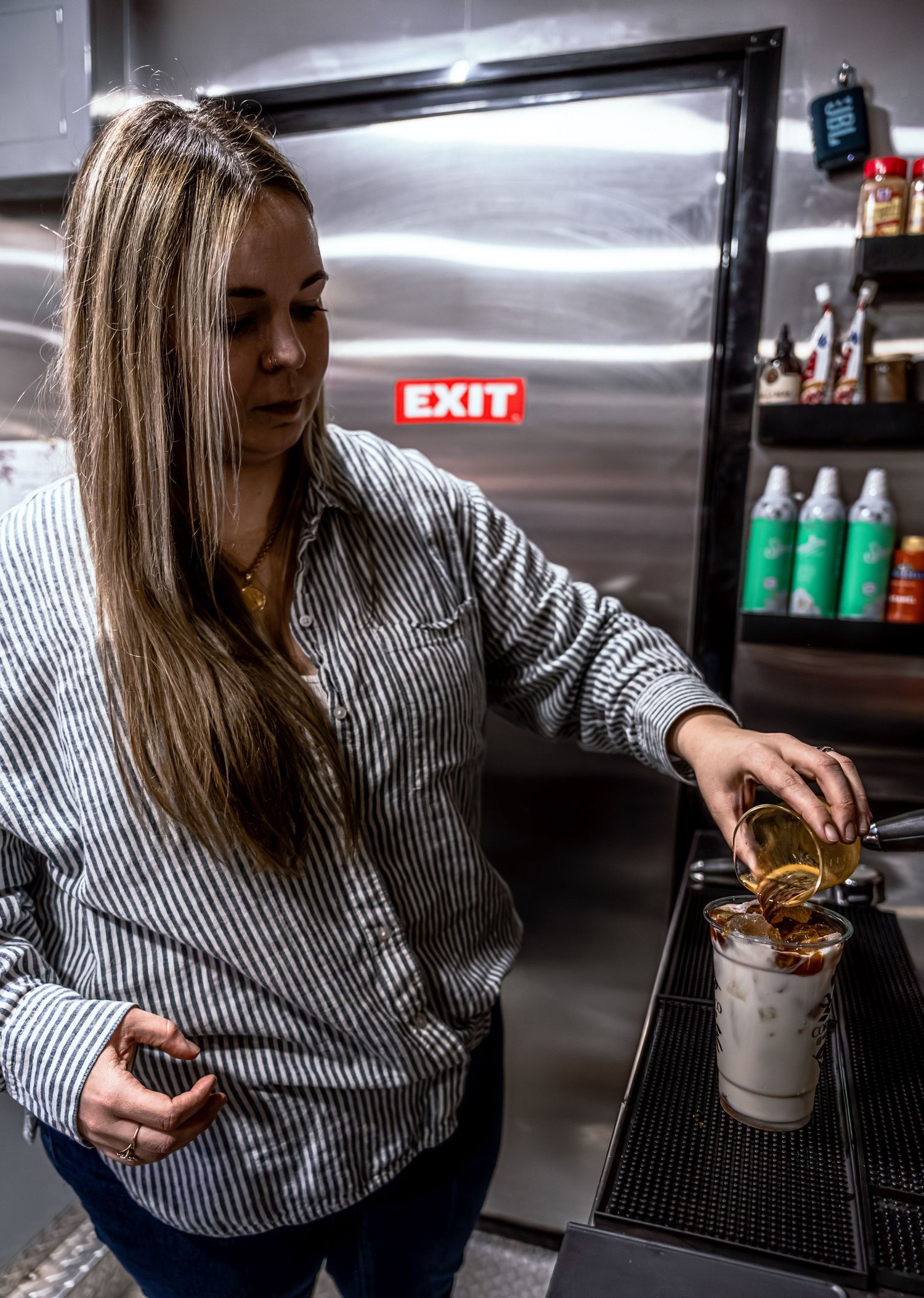 Person in striped shirt preparing coffee behind counter in dimly lit space.