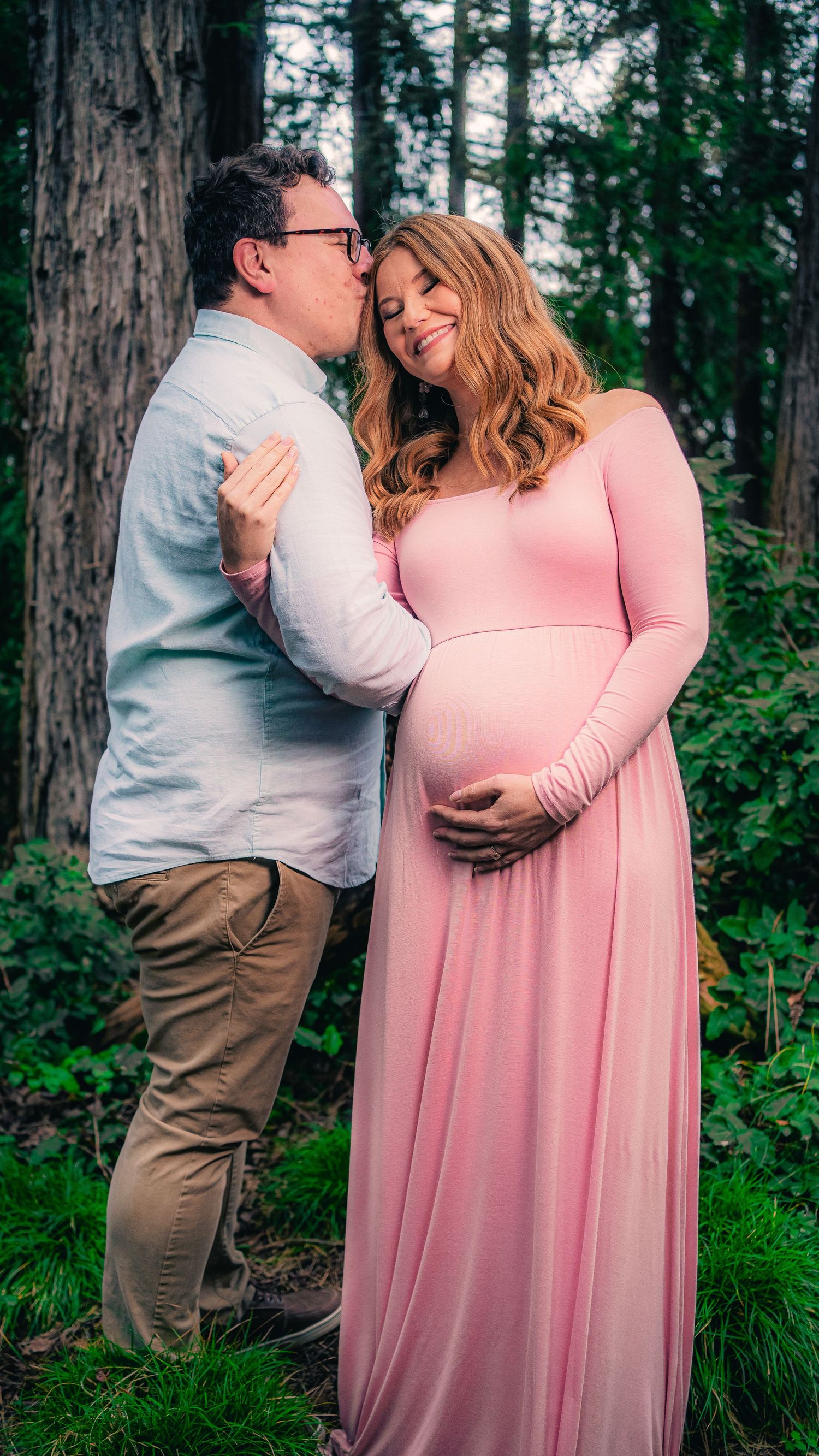 Couple embraces in a forest setting wearing pink dress and white shirt against tall trees in background.