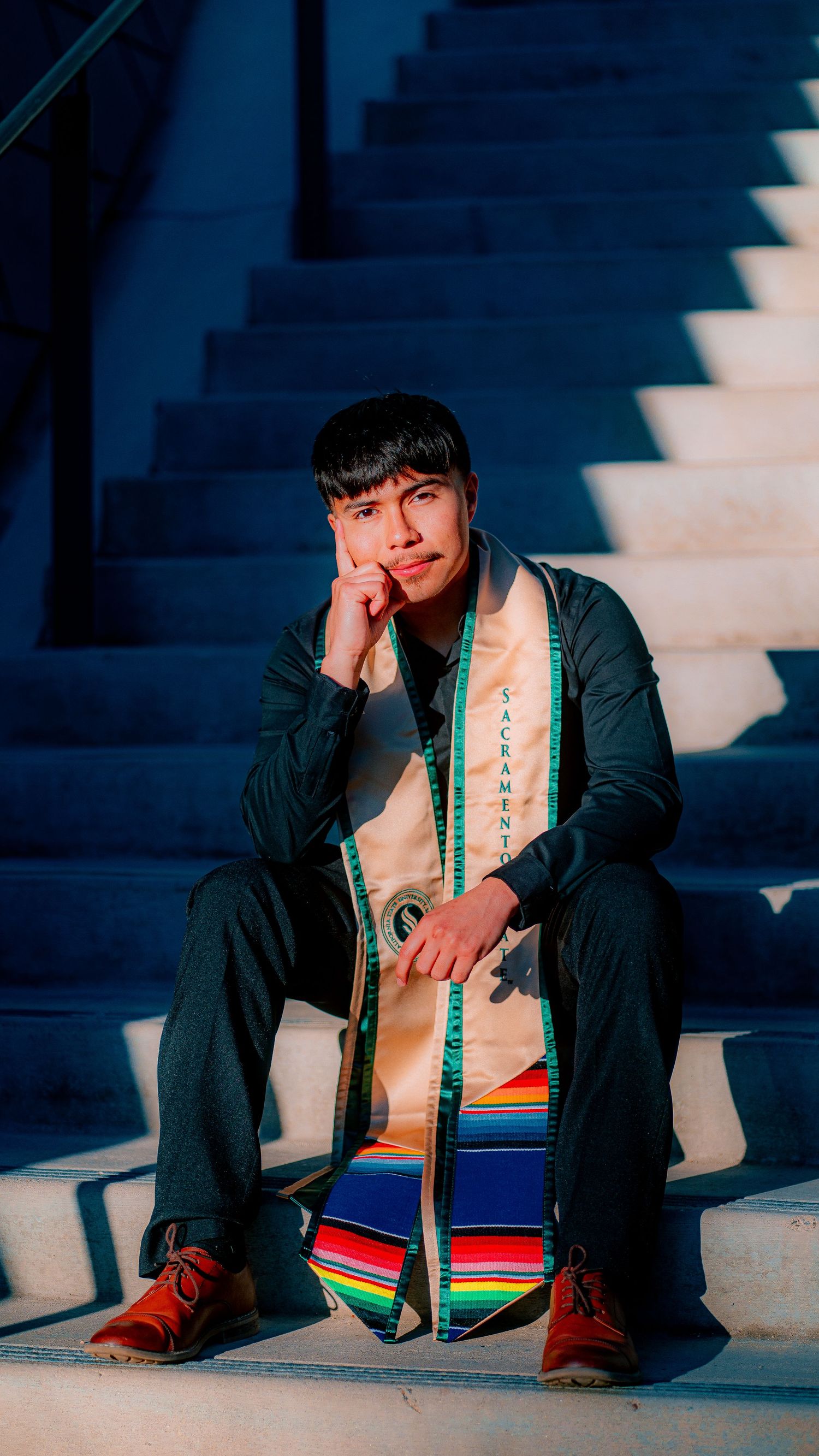 Graduate in black gown and colorful stole sits on concrete steps in dramatic lighting.
