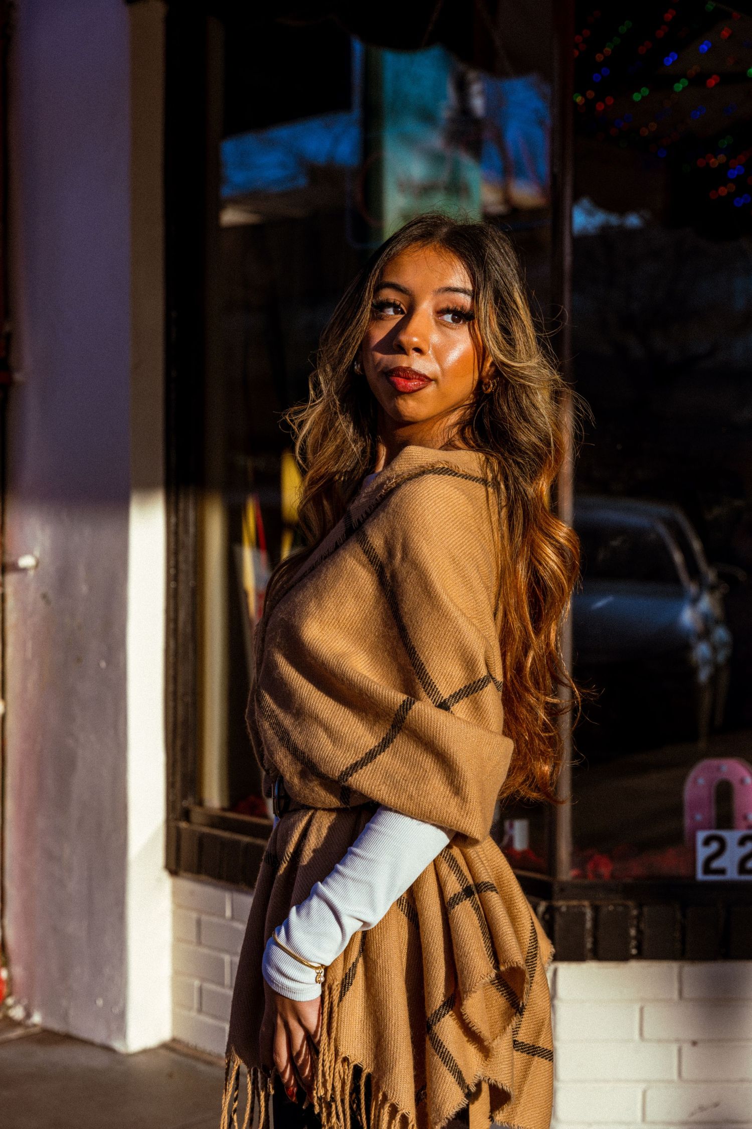 Fashion portrait against white brick wall featuring tan fringed wrap and white top.