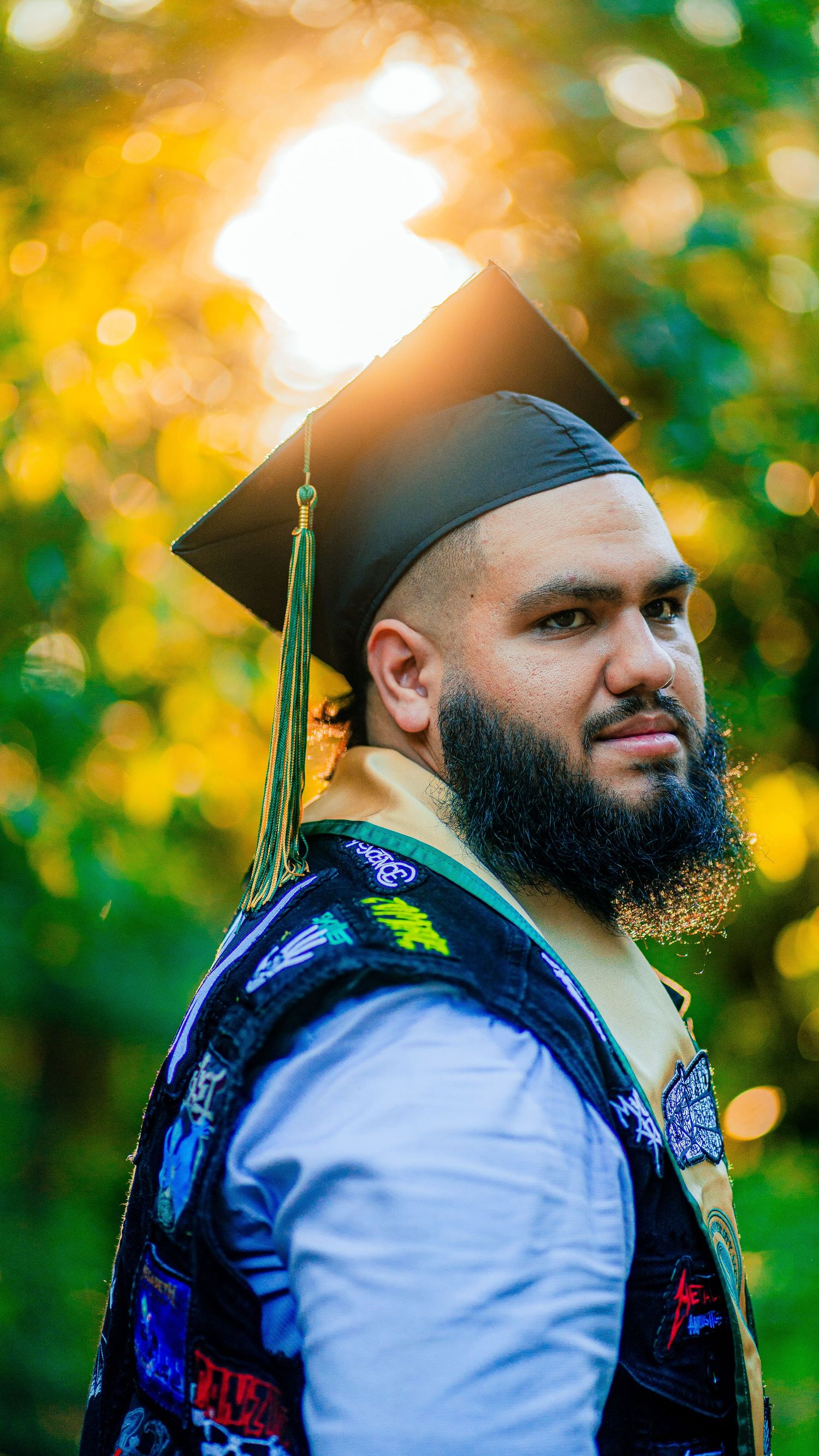 Graduate in cap and gown poses against blurred autumn background with bokeh lighting effects.