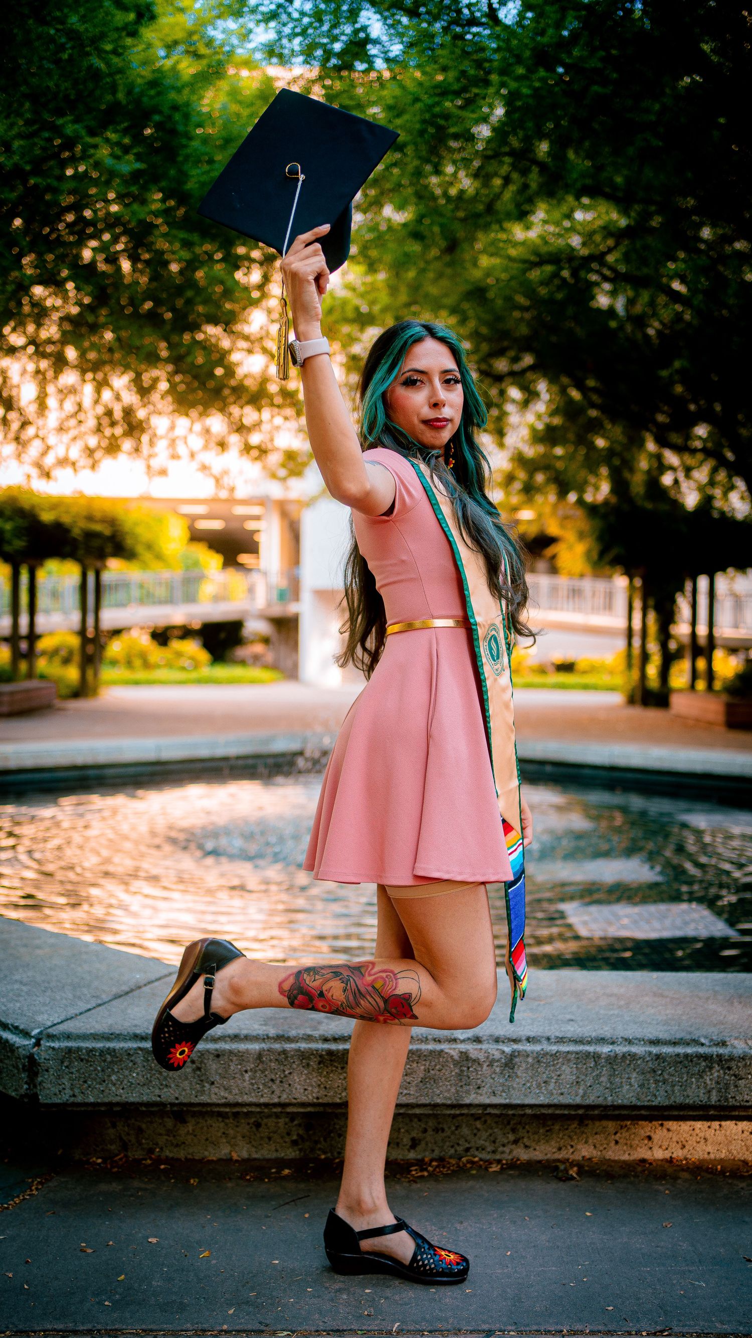 Graduate in pink dress striking playful poses on campus grounds.