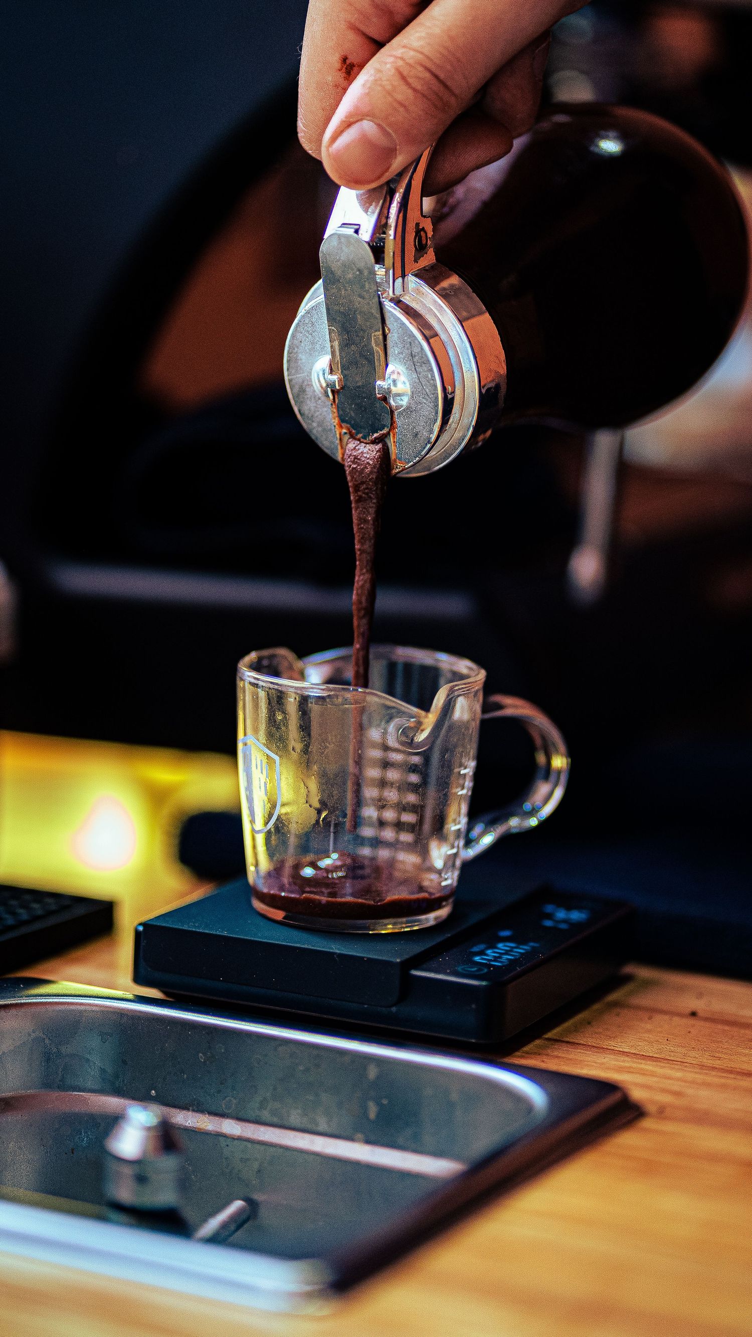 Hands carefully pouring espresso shots into measuring cups on a wooden surface.