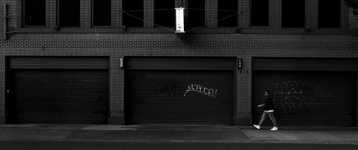 A lone pedestrian walks past dark storefronts on an empty street illuminated by a single light in black and white.