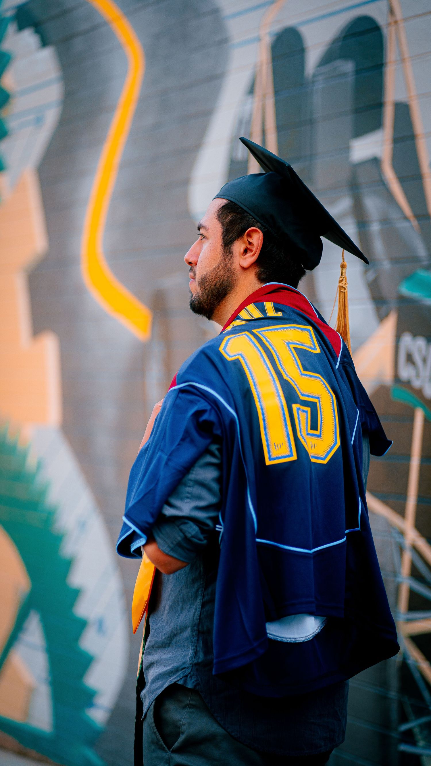Graduate wearing blue and yellow graduation stole and mortarboard cap stands against abstract background.