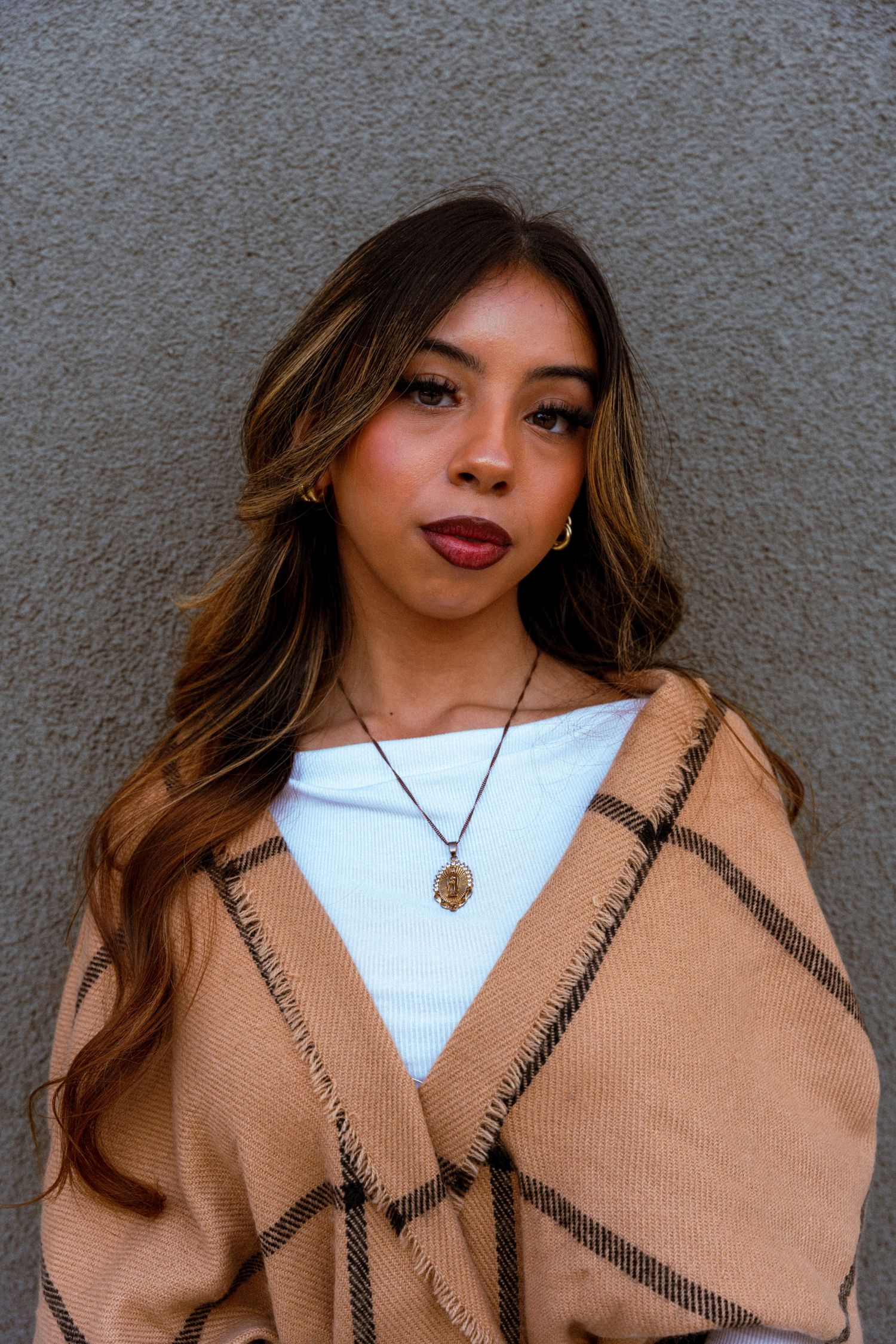 Fashion photo showing a camel-colored shawl with black trim styled over a white top against a gray wall.