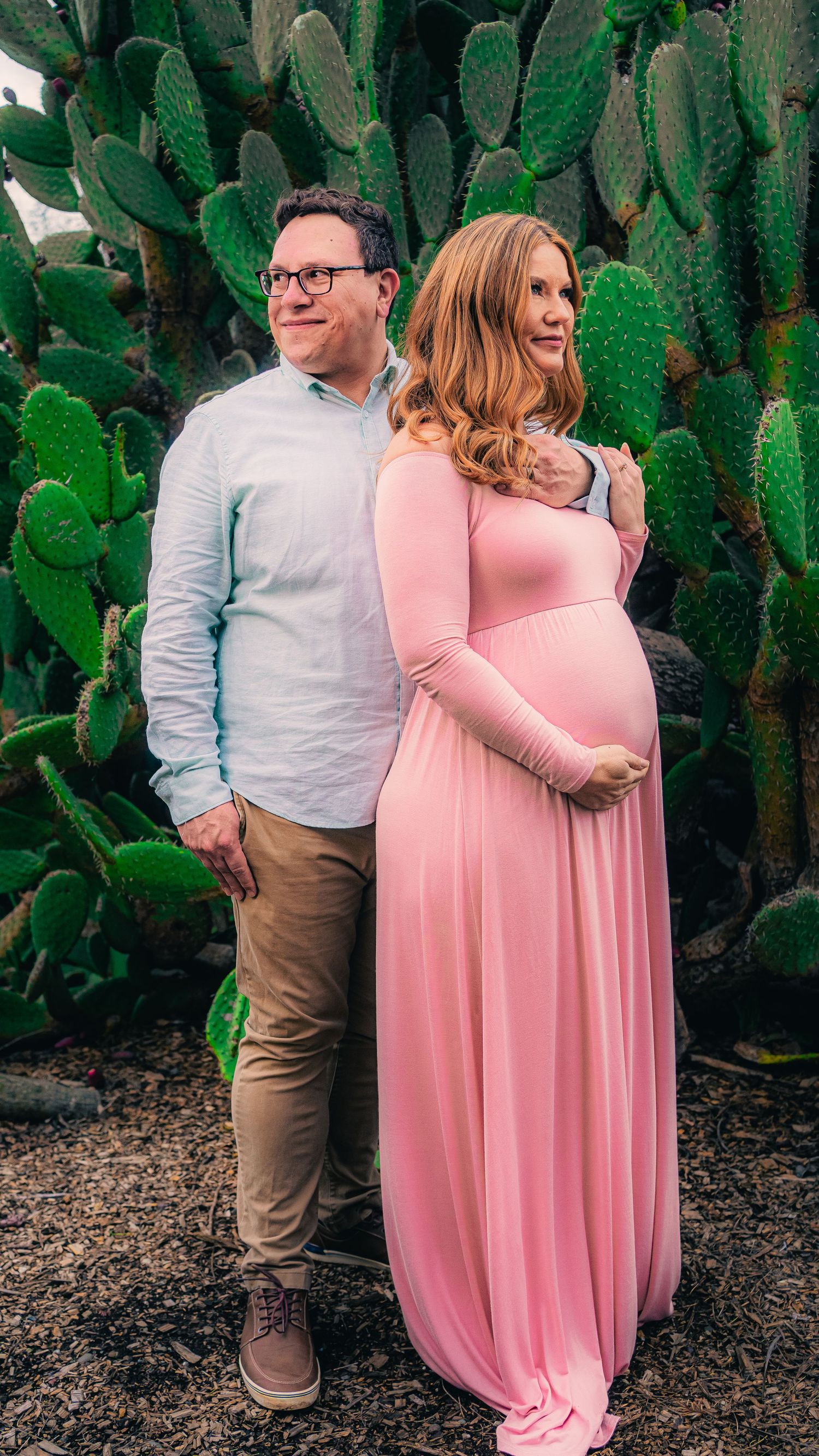 Sweet couple poses together by desert cactus garden wearing pink dress and white shirt.