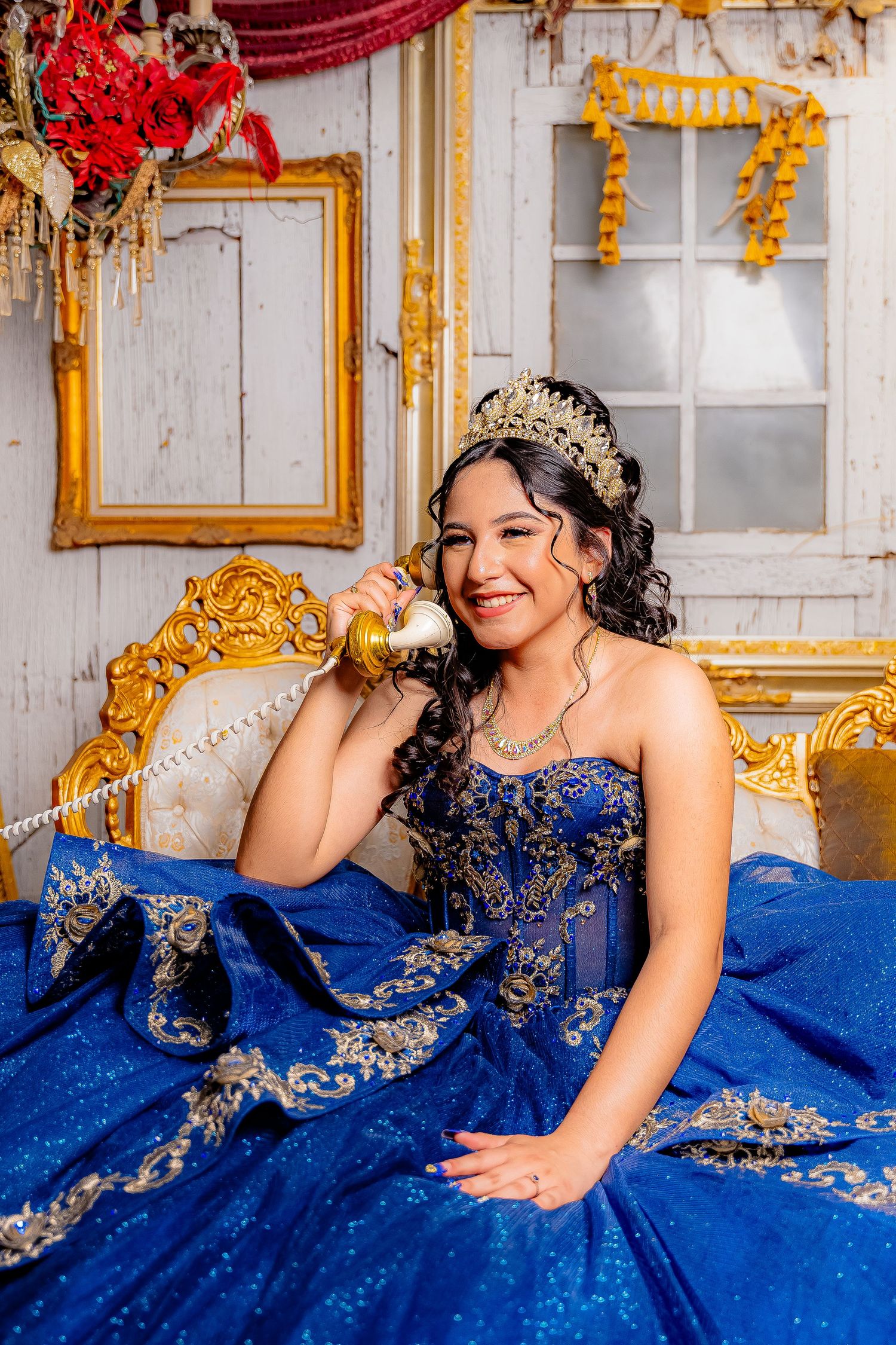 A quinceañera celebration featuring a person in a royal blue ballgown with gold embellishments posing on a luxurious sofa.