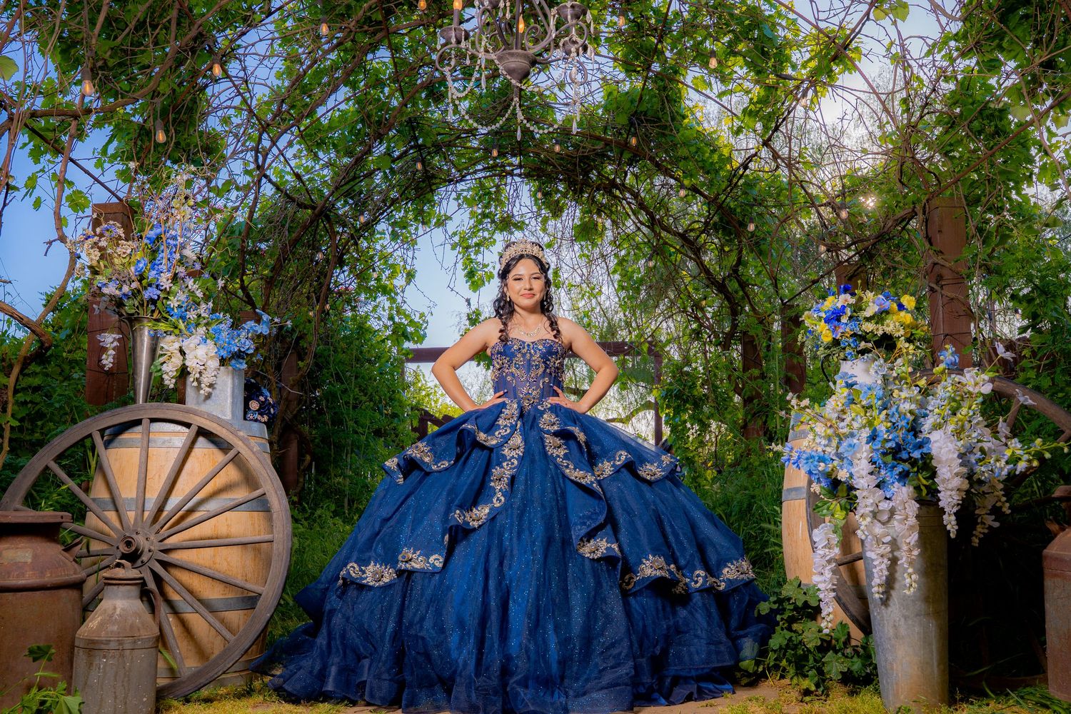 Majestic quinceañera photo featuring a blue ballgown in a garden setting with rustic wagon wheel.