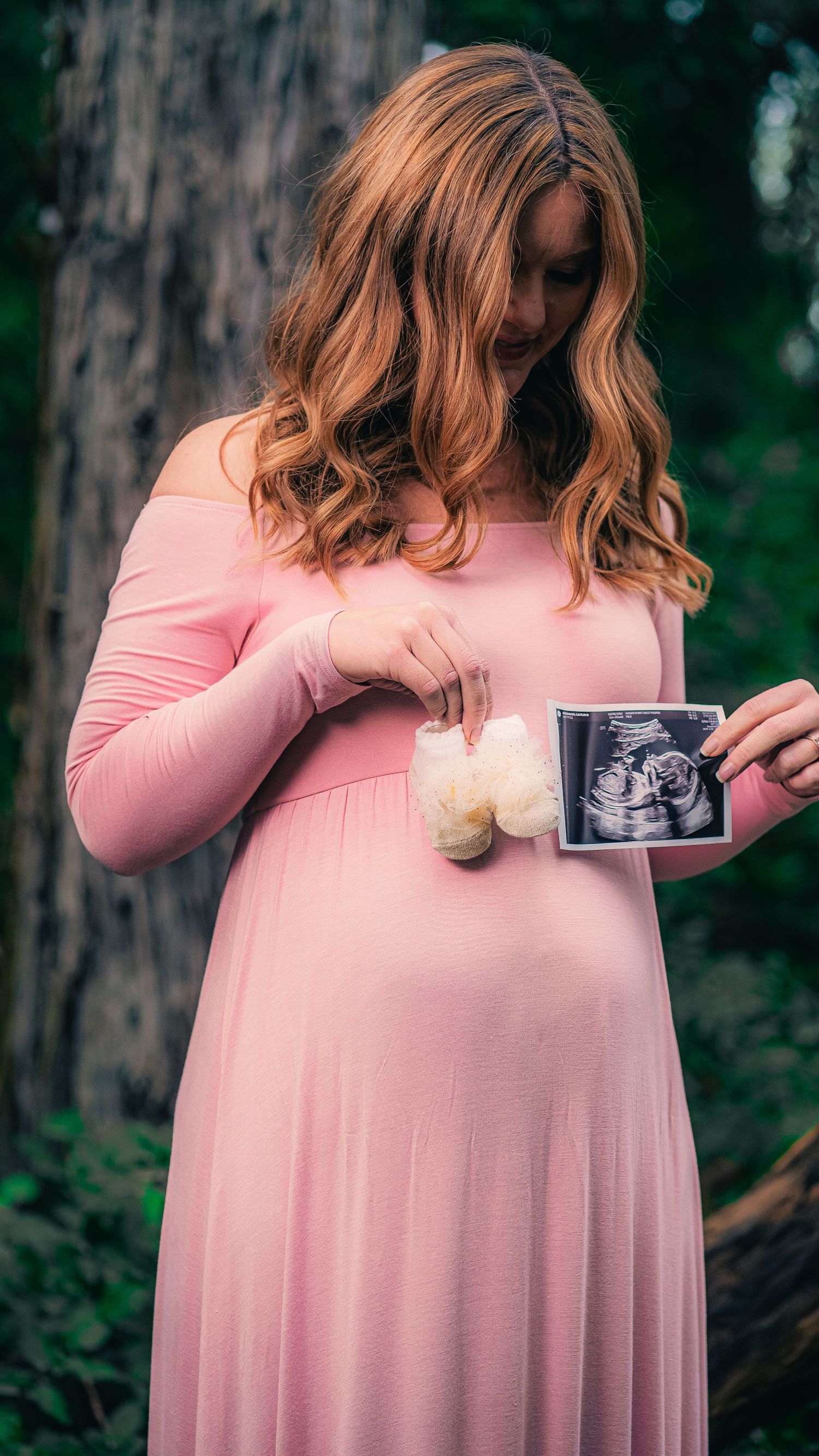 Expectant mother in pink dress holds ultrasound photo while standing in forest setting.