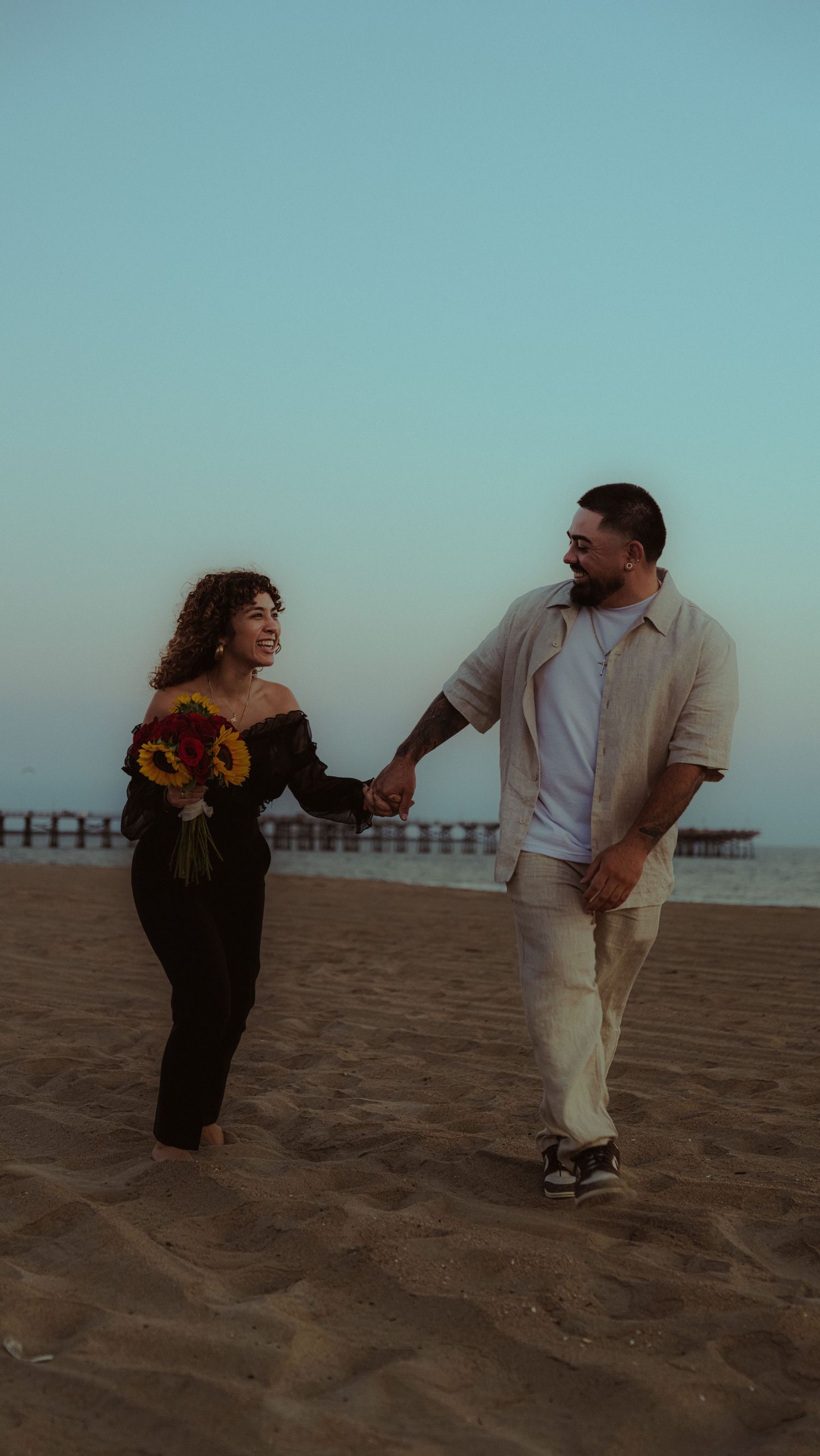Playful couple running and dancing on a sandy beach with sunflowers.