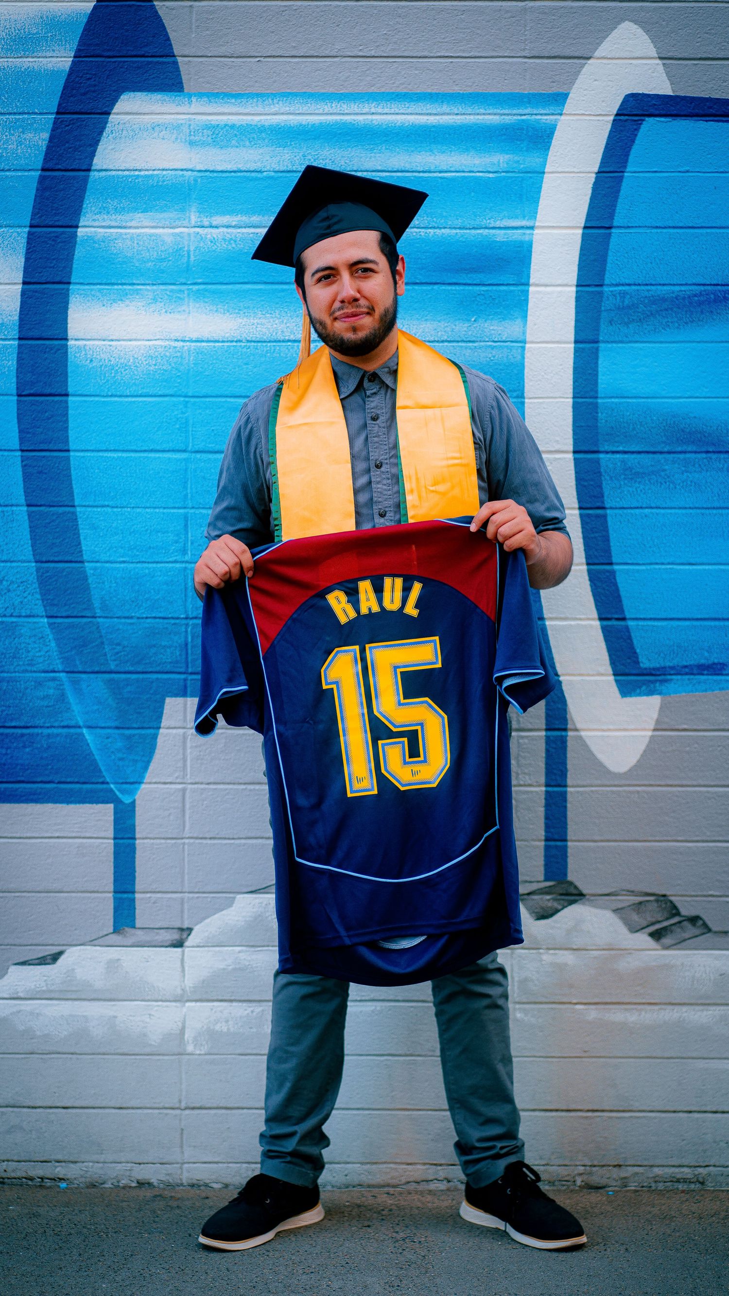 Graduate displaying sports jerseys with numbers against blue wall backdrop.