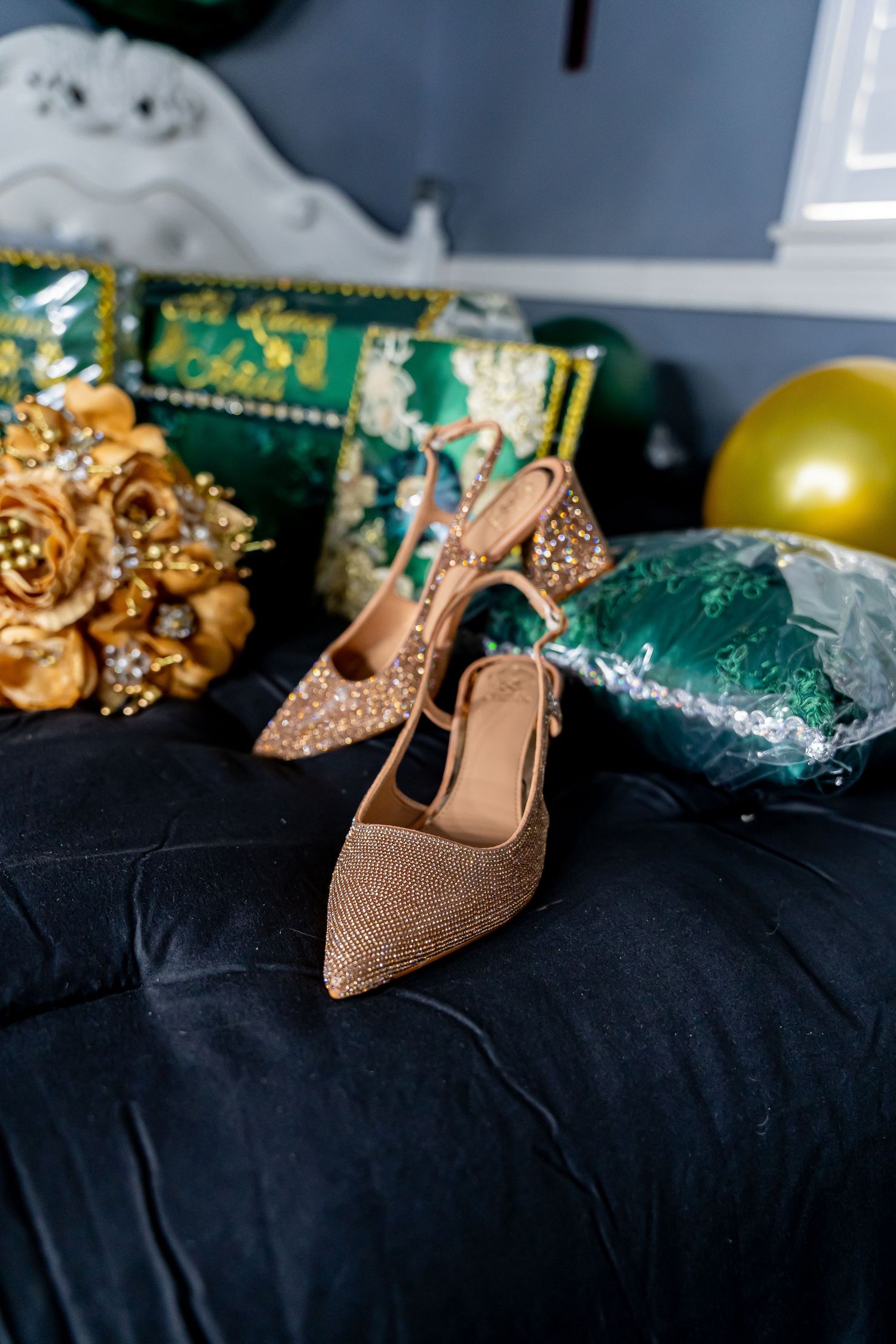 Sparkly gold high heels and accessories arranged on dark fabric with emerald green decorative elements.