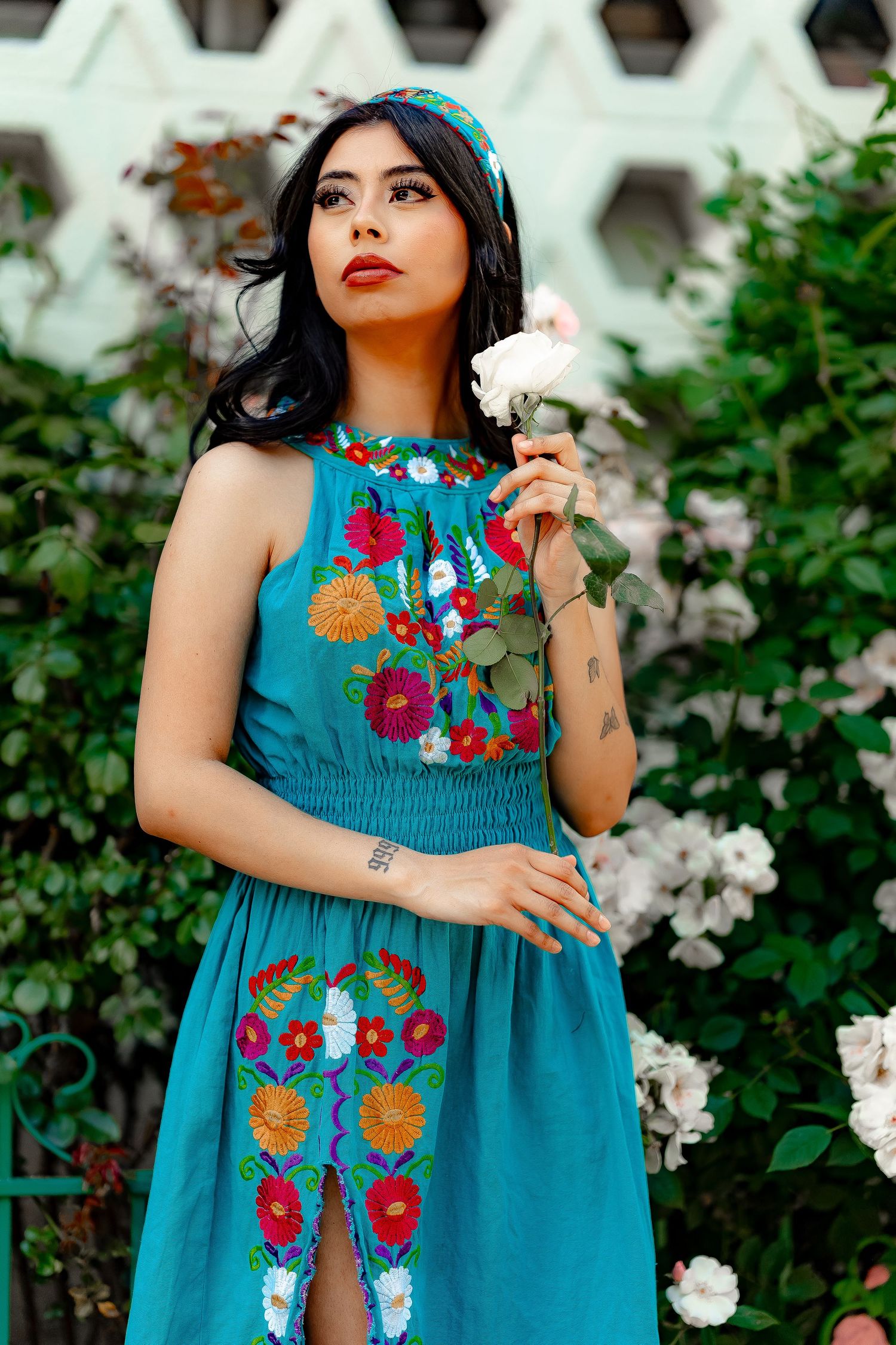 A teal sundress with vibrant floral embroidery details is showcased in a garden setting with white roses.