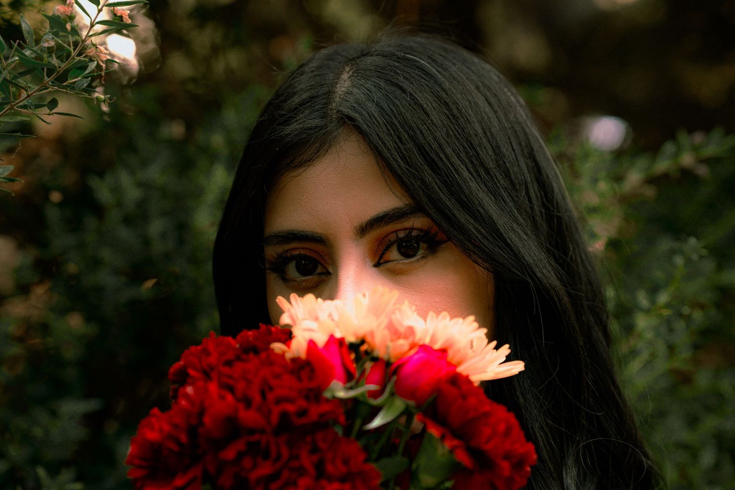 A person holds vibrant red carnations in front of their face against a lush garden background.