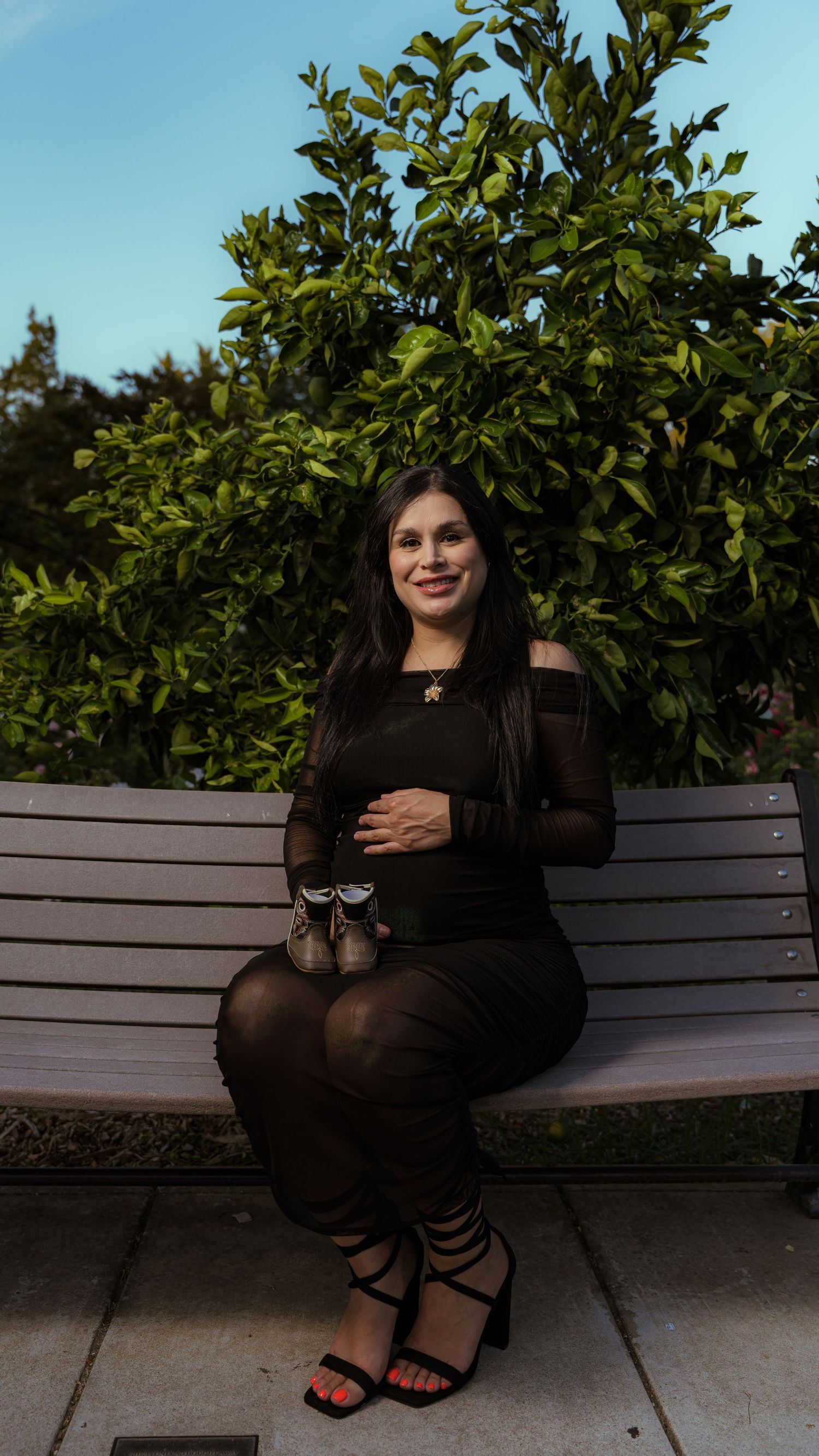 A maternity portrait on a wooden bench against lush green foliage.
