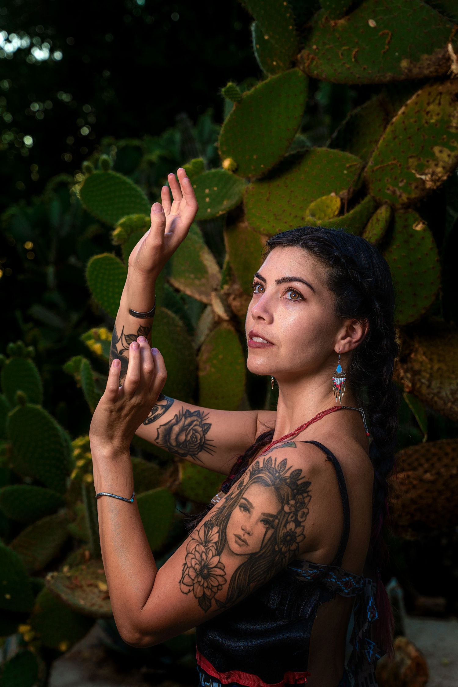 An artistic pose against cactus plants showcasing intricate tattoo art and a black dress with red details.