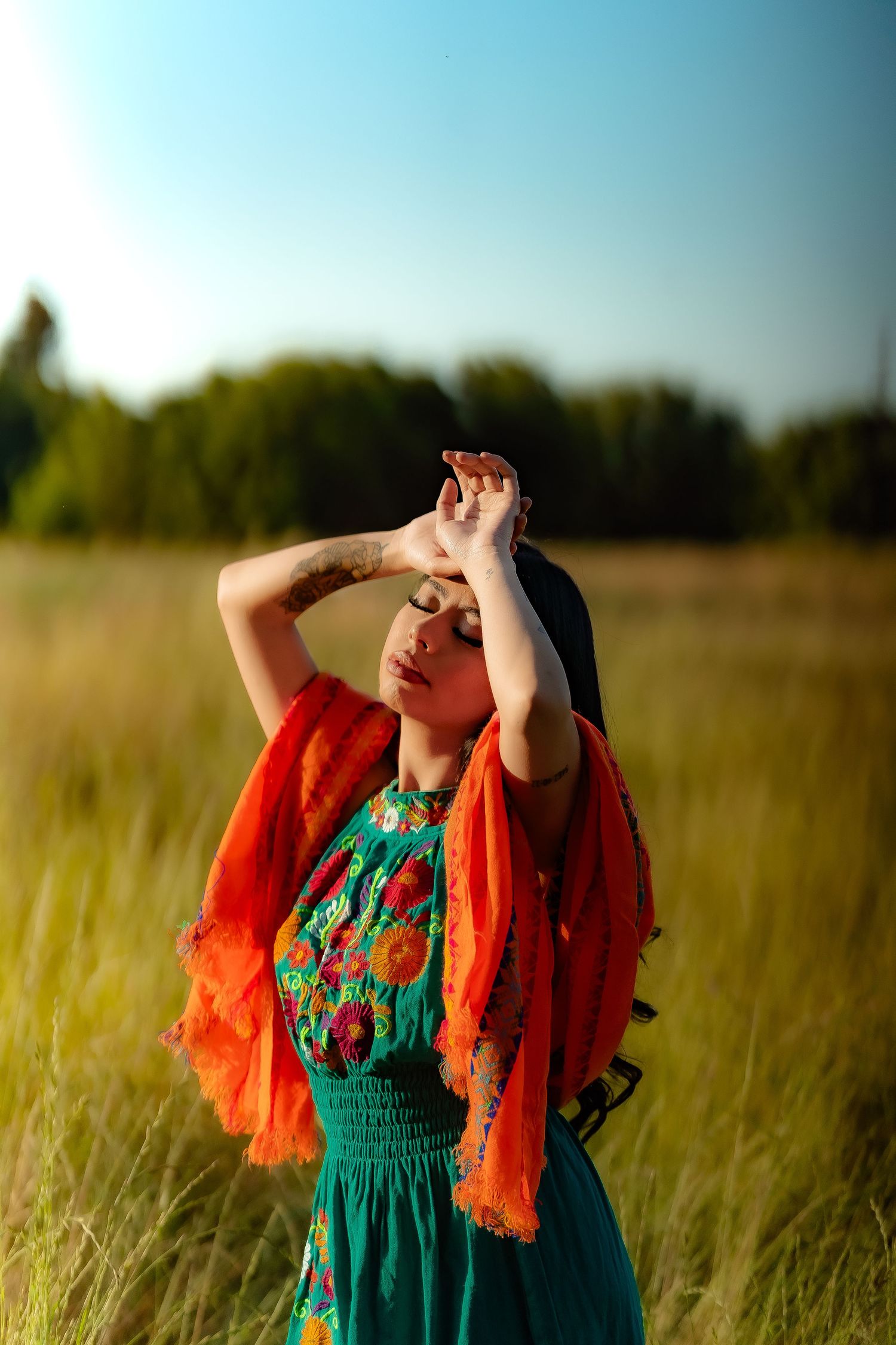 A flowing orange shawl and teal dress create movement against tall grass in a dreamy outdoor portrait.