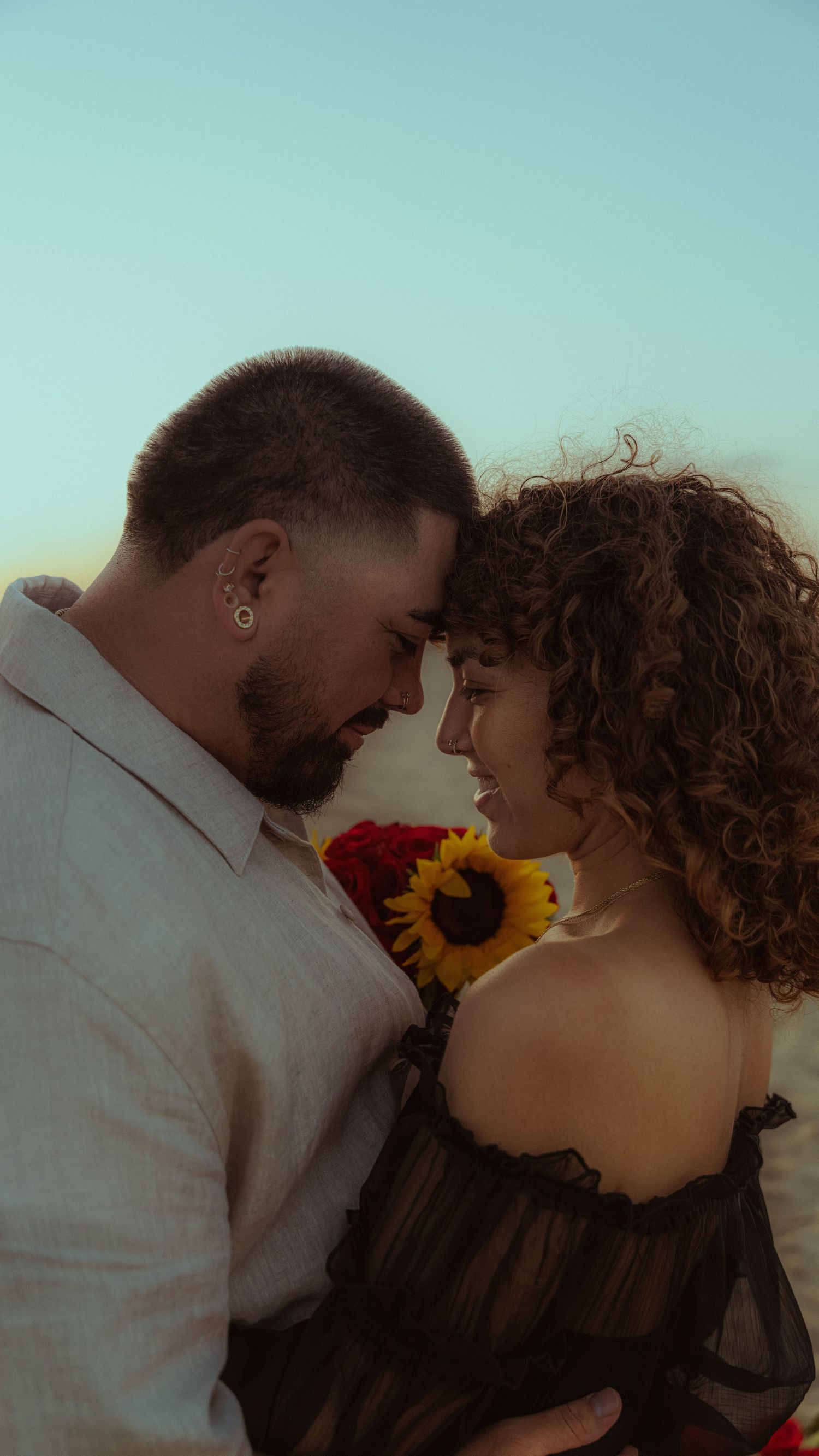 A couple shares tender moments with sunflowers against a blue sky background.