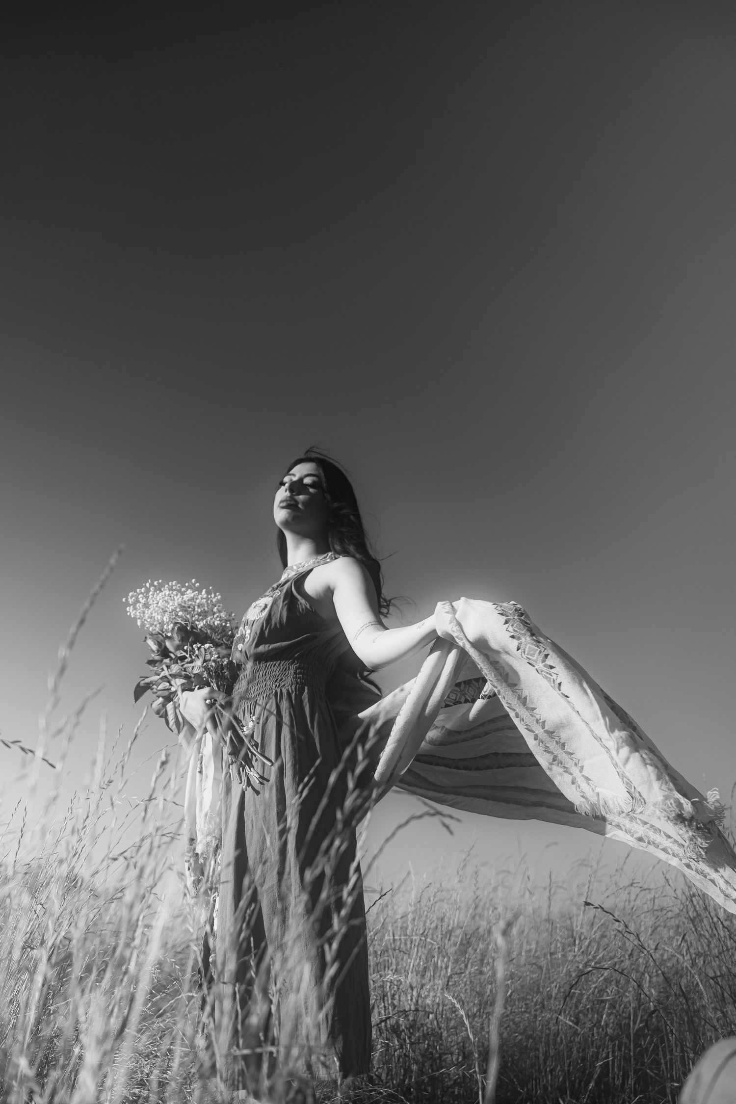 A black and white artistic photo of a figure in a flowing dress holding flowers while standing in a grassy field.