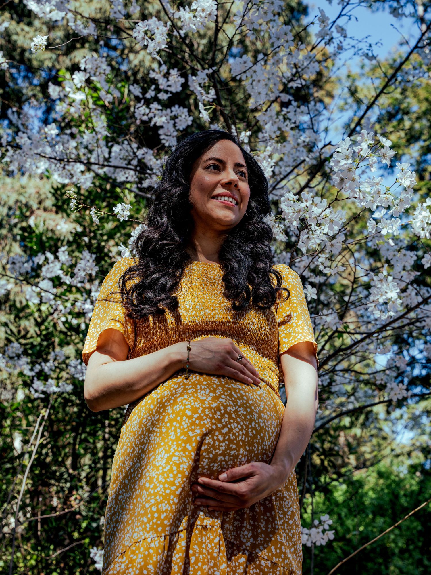 Expectant mother in yellow floral dress poses beneath blooming cherry blossom trees on a sunny spring day.
