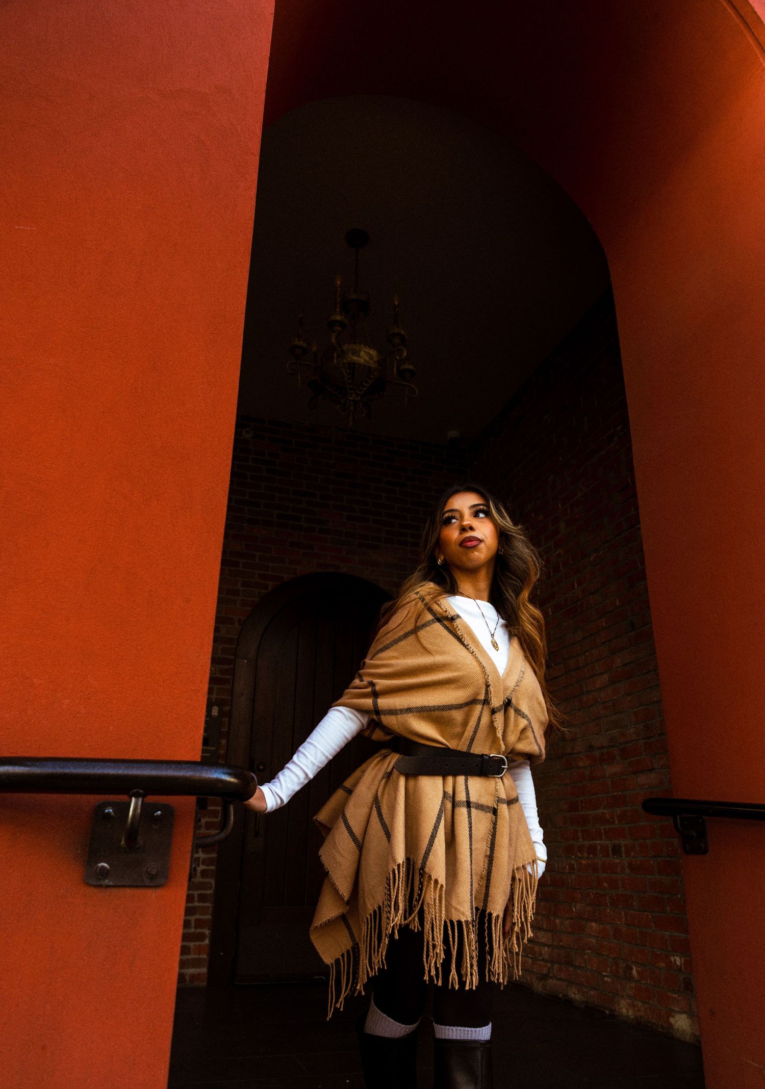 Figure wearing a camel-colored fringed wrap poses dramatically against a vibrant red wall.