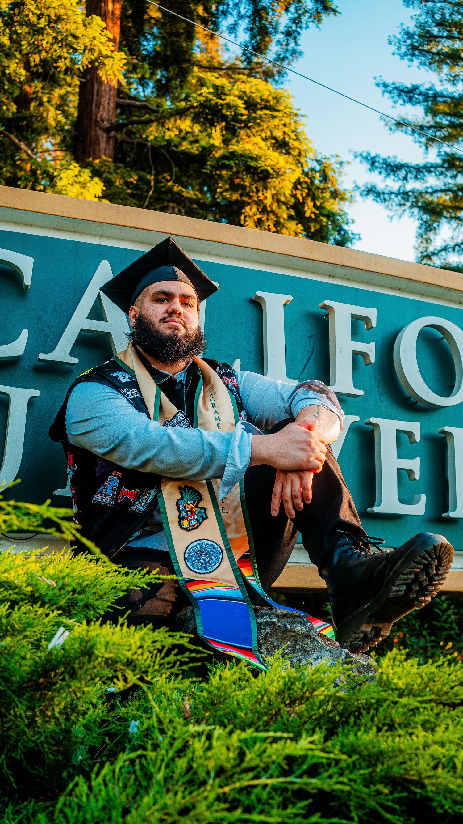 Graduate in blue regalia sits proudly by university signage.