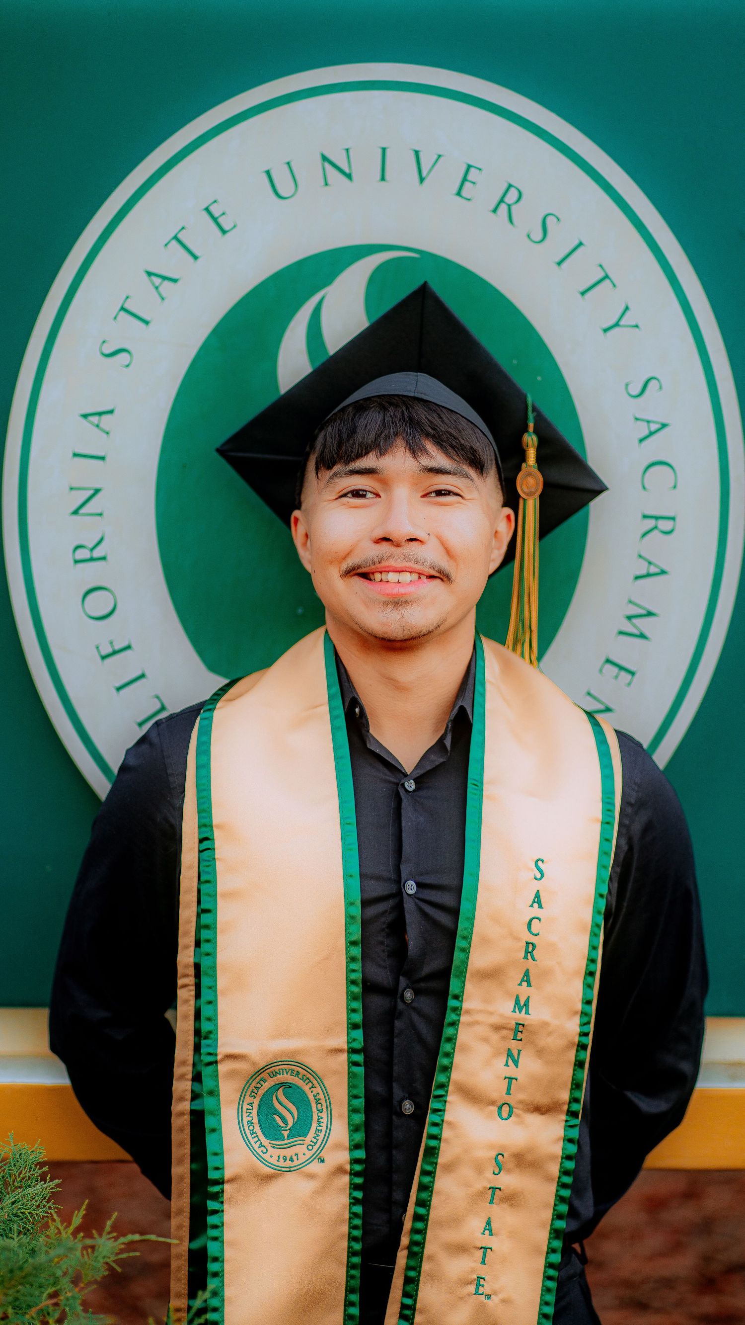 Graduate wearing green and gold stole poses in front of university logo backdrop.
