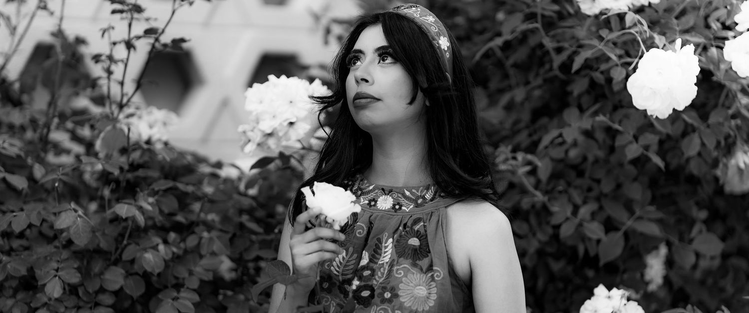 A woman in a white dress poses elegantly among lush greenery and white flowers in a garden setting.