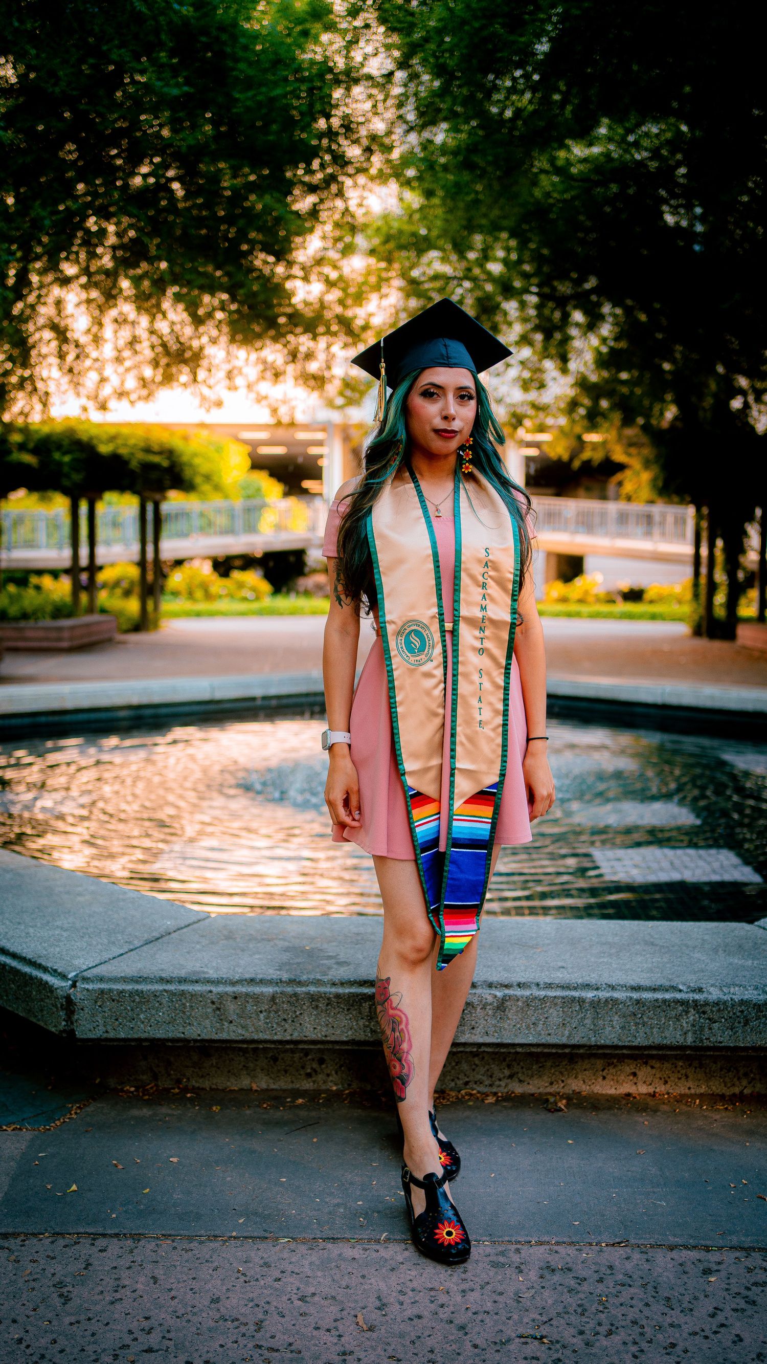 Graduate in pink dress and cap posing on campus pathway at sunset.