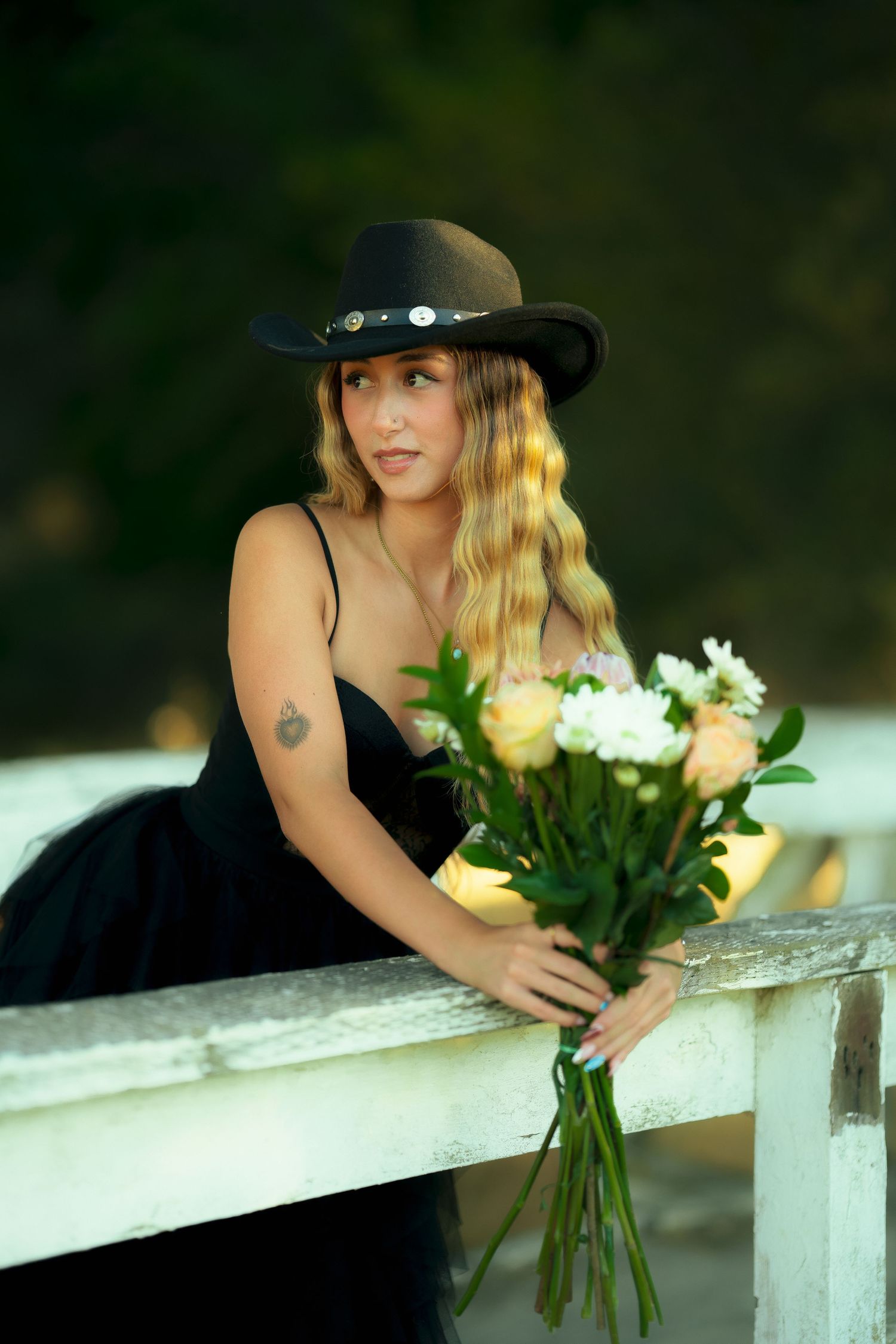 A person in a black dress and cowboy hat poses with yellow roses on a wooden fence.