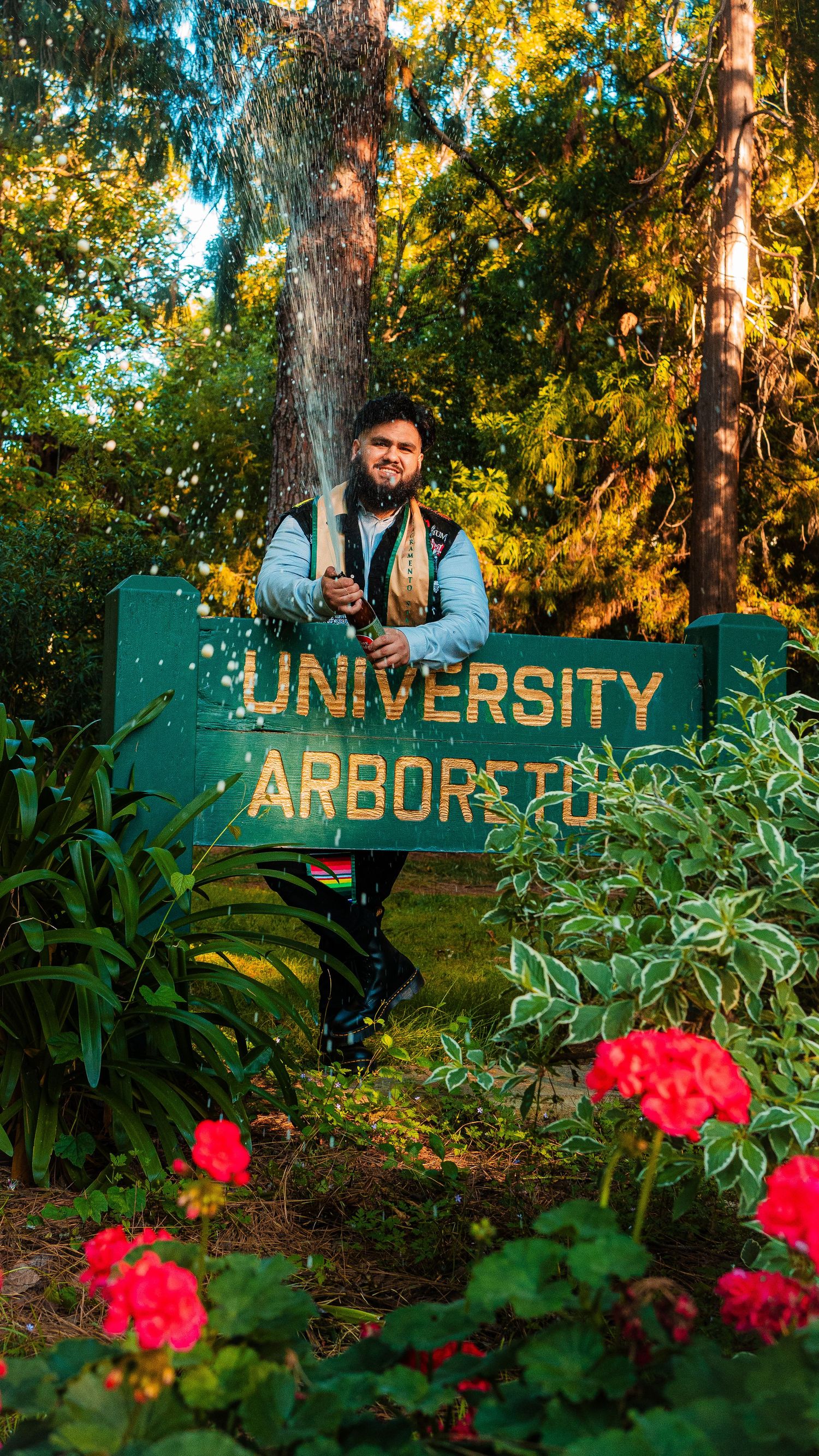 Graduate poses at University Arbors sign surrounded by flowers and trees.