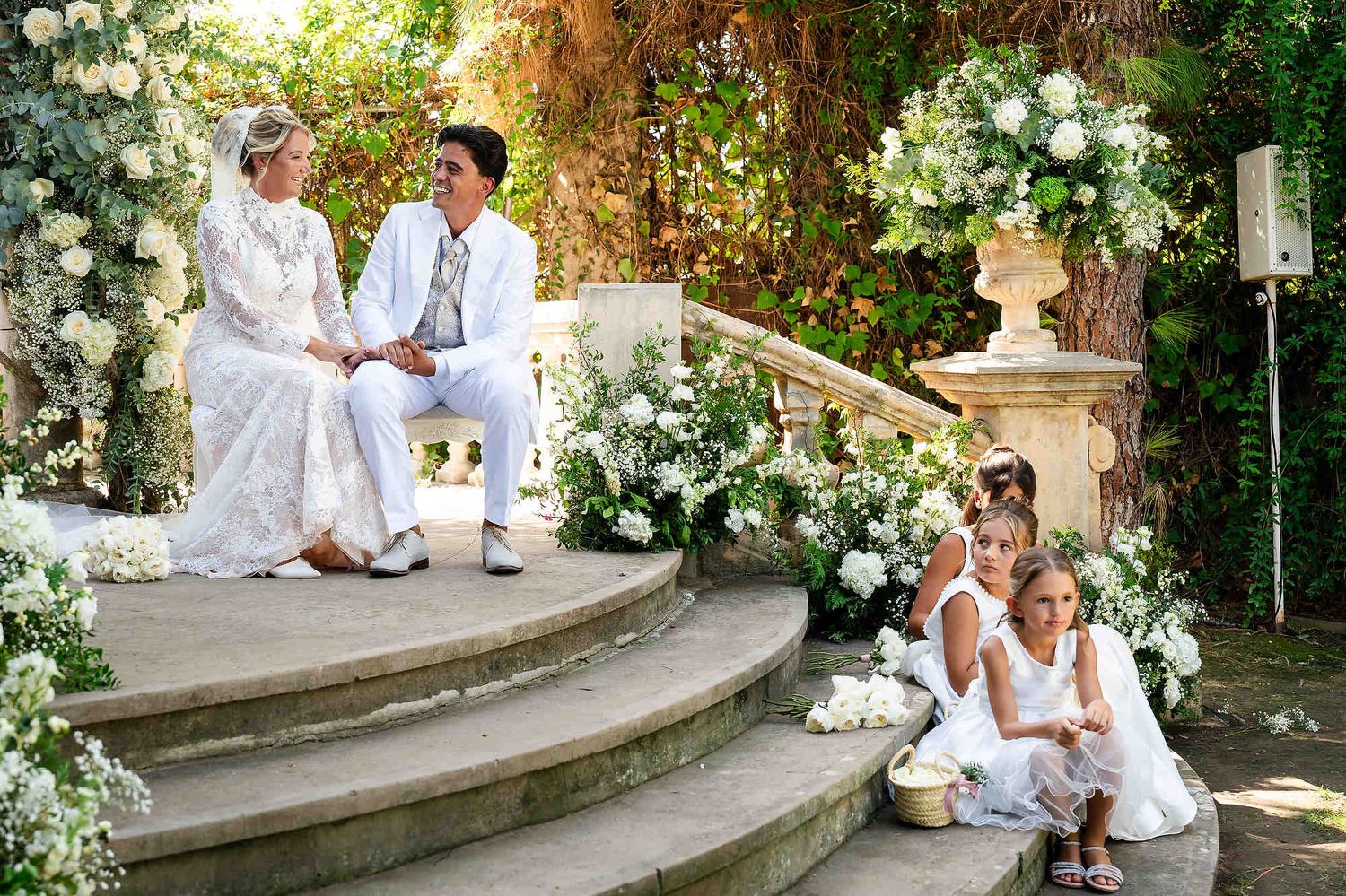 The bride and groom seated at the antique stone ceremony arch with the bridal party in formal attire on the steps.