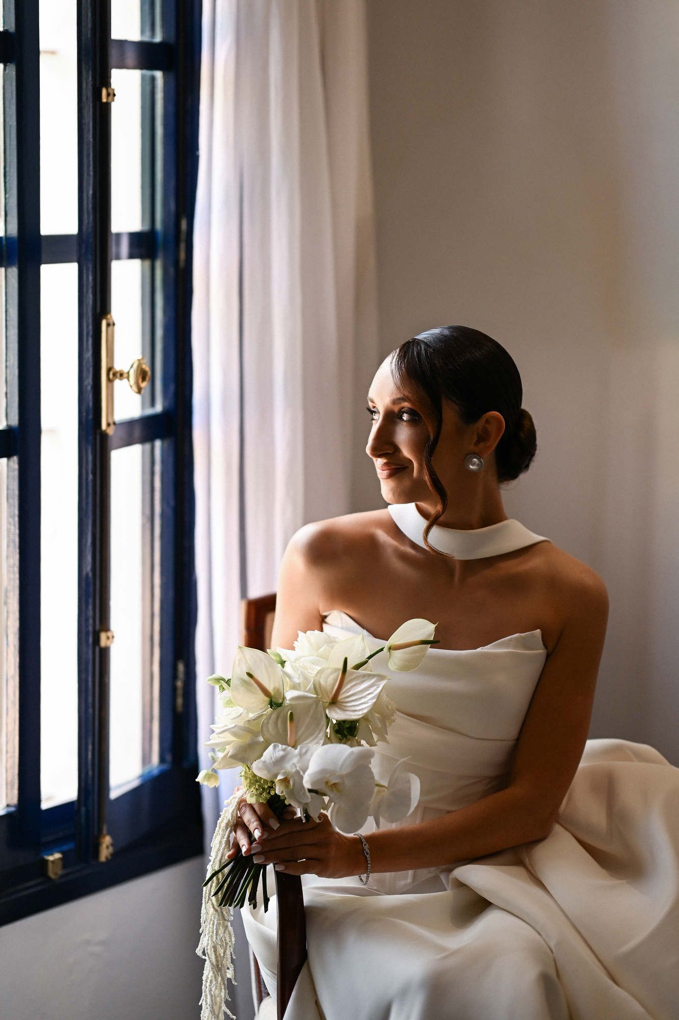 Bride with elegant hair and makeup looks out the Villa Paula window holding a calla lily bouquet before the ceremony in Spain.