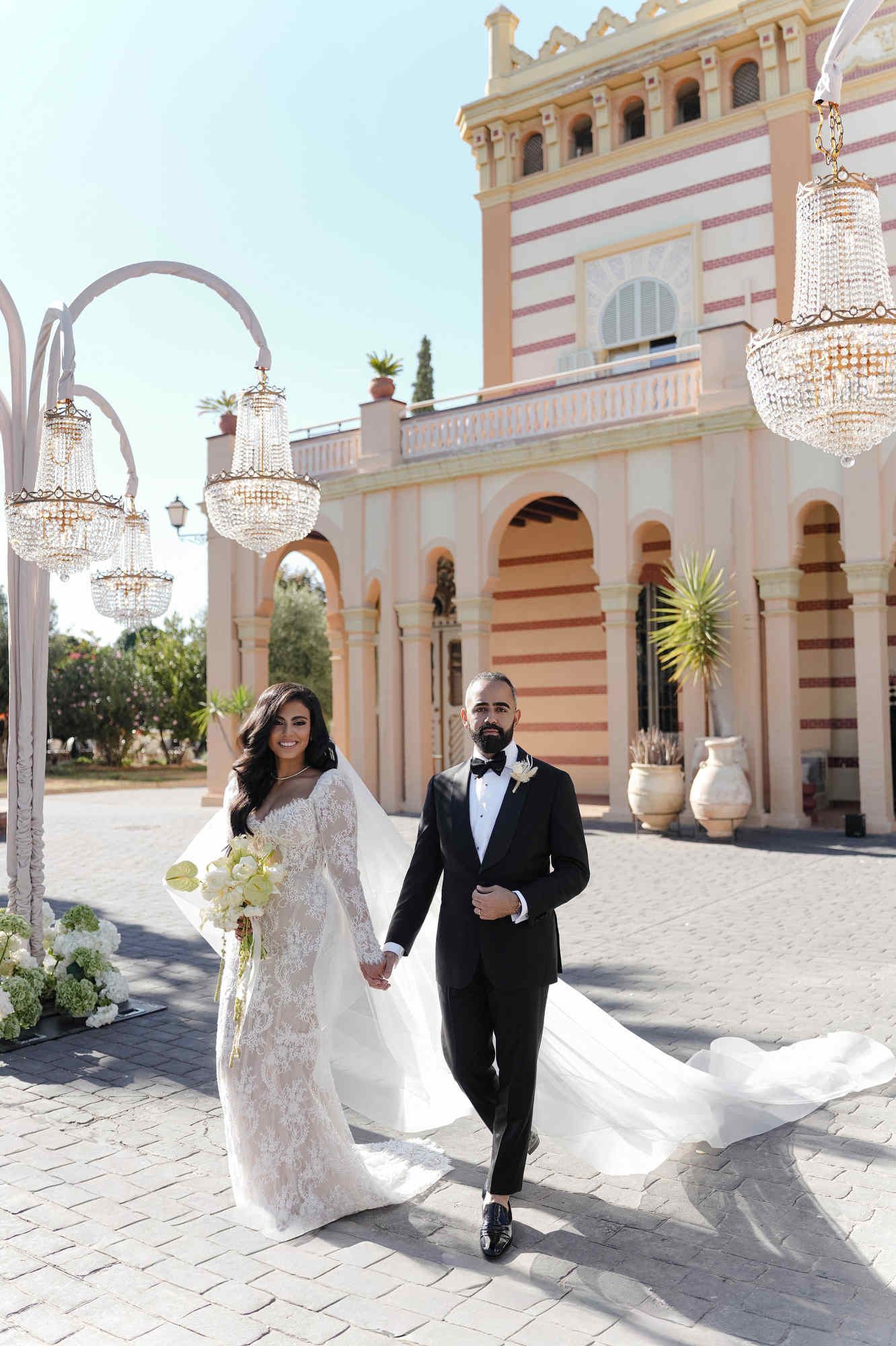 The couple walking to their ceremony at Gran Villa Rosa; bride with a long train and groom in a classic black suit.