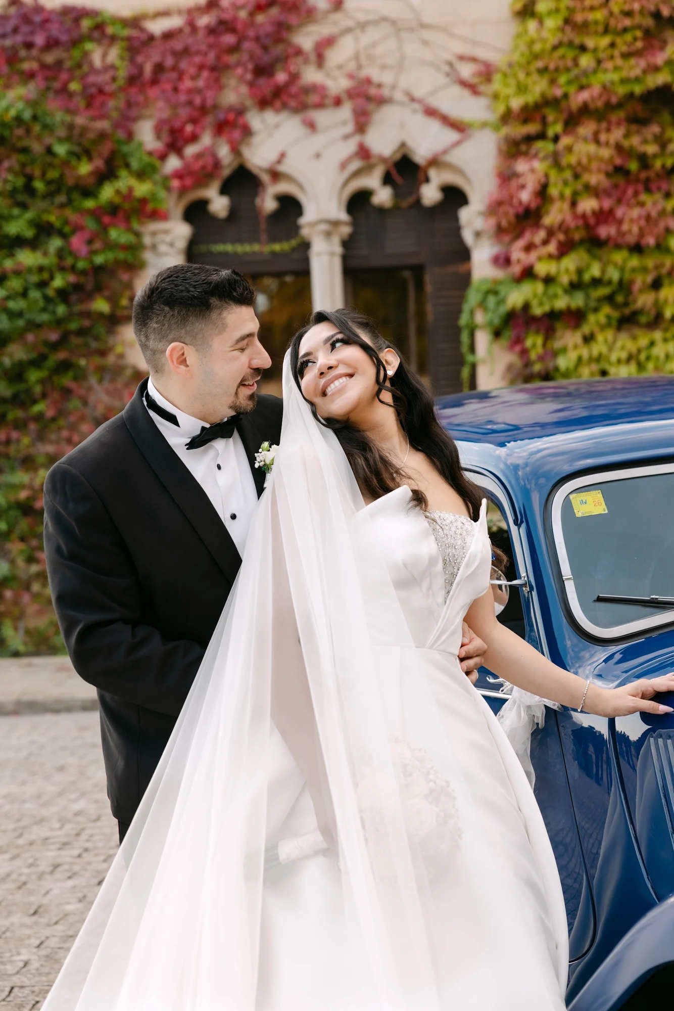 The newlyweds posing with a stylish blue retro car in front of the historic stone facade of Castell Jalpí