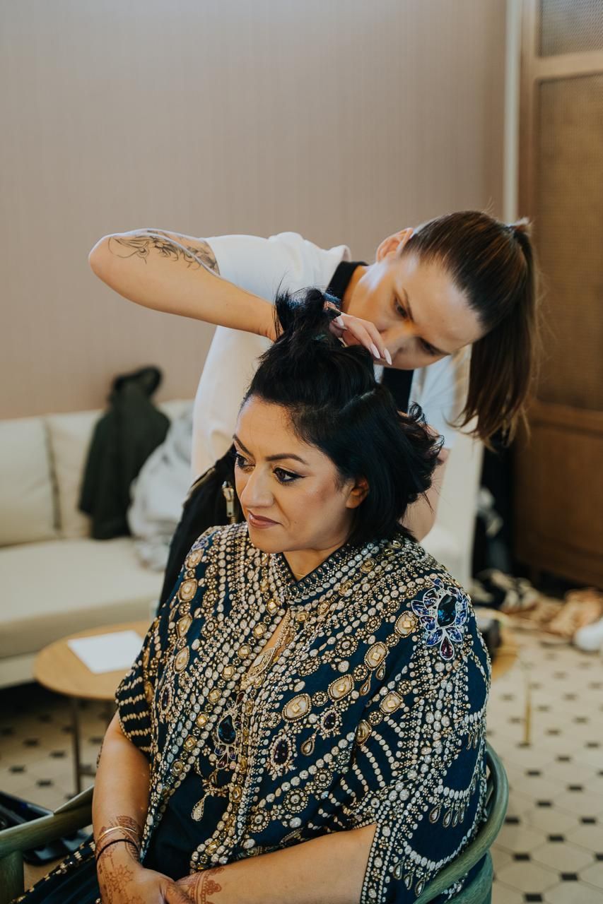 The makeup artist intricately styling the hair of the second Indian bride during their joint wedding preparation.