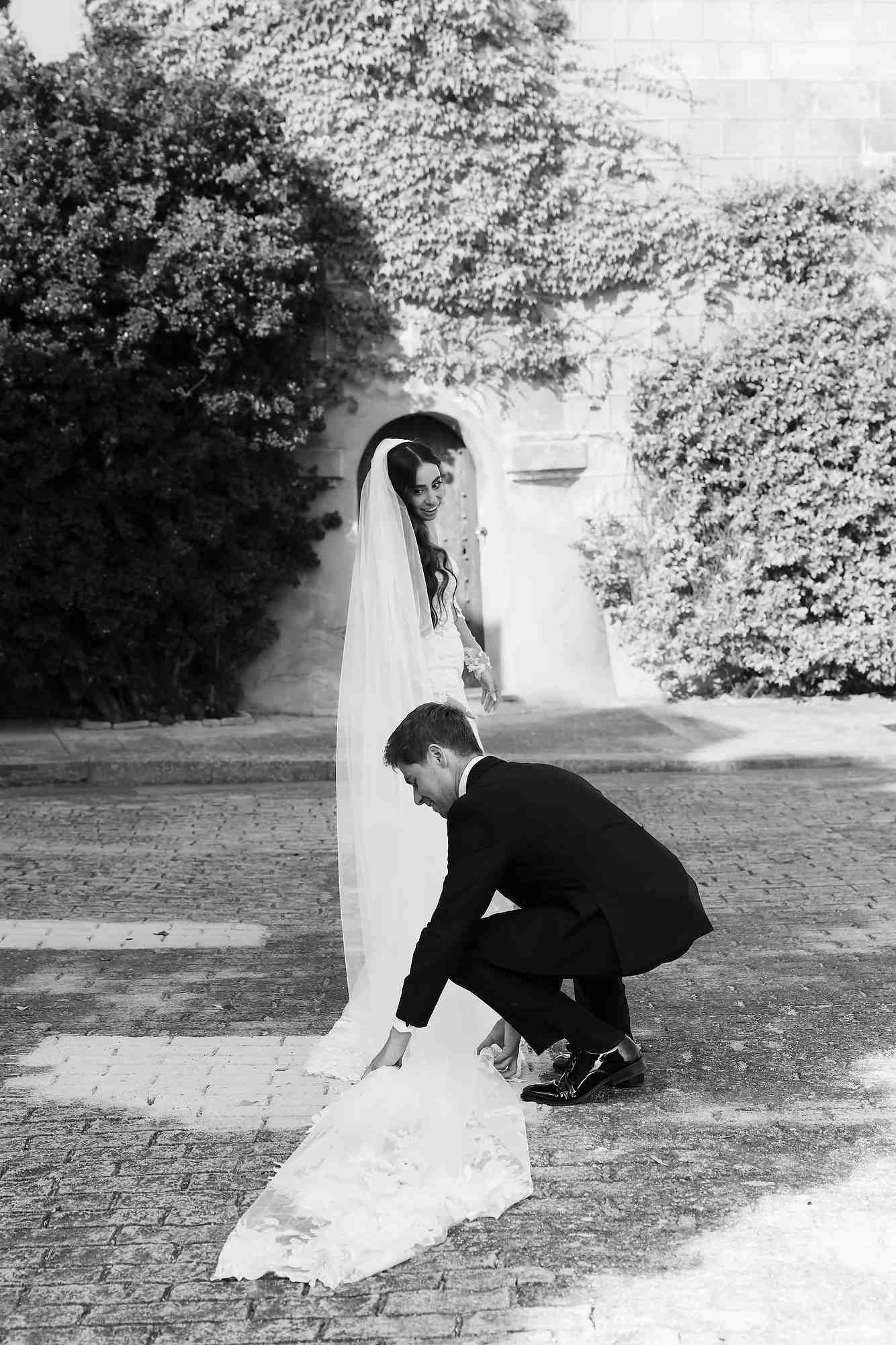 A candid moment of the groom fixing the bride’s train during their photoshoot, showing the resilience of her hair and makeup.