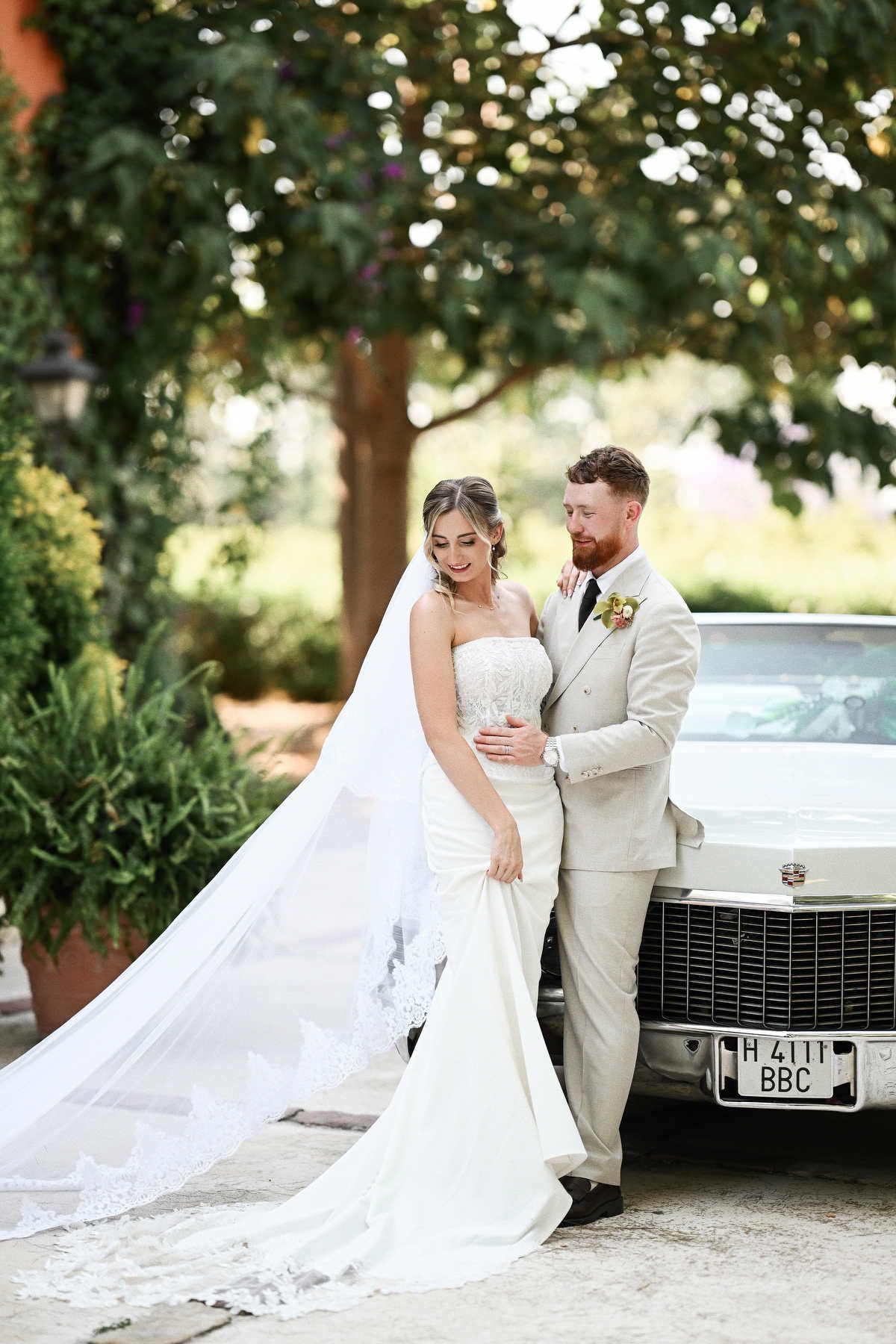 Bride and groom posing with a vintage car. Long bridal veil professionally secured into her wedding hairstyle by a luxury hair stylist.