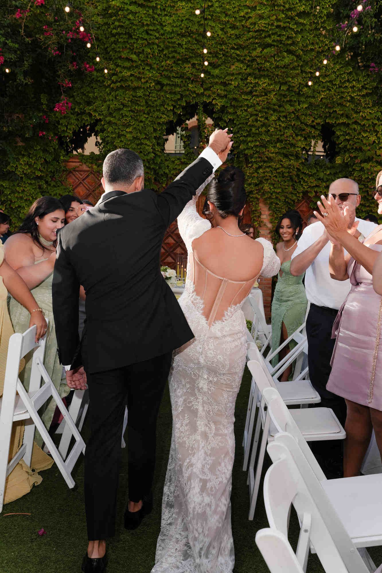Newlyweds walking to guests; the bride’s open-back dress highlights her elegant bridal hair styling at Gran Villa Rosa