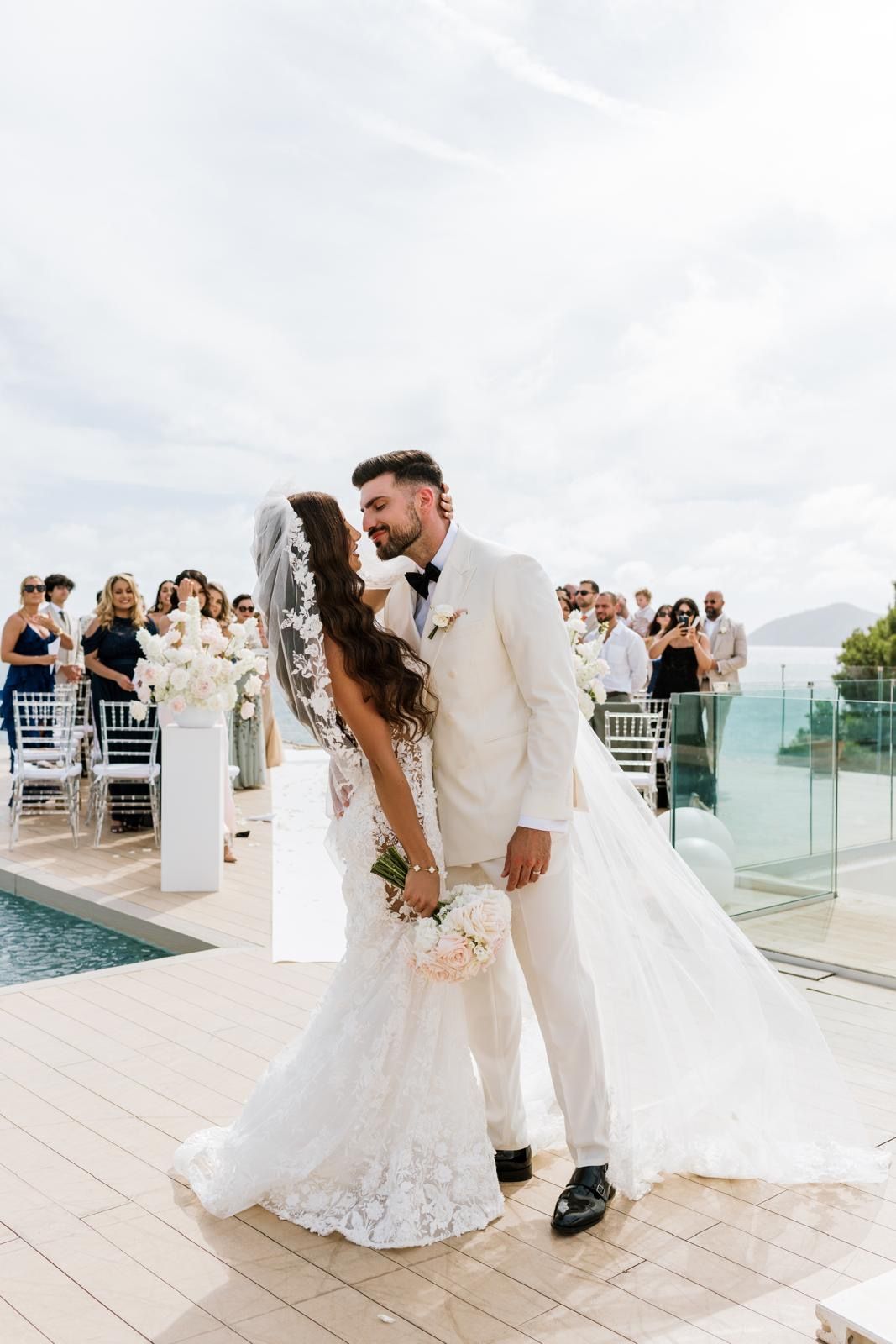 The newlyweds sharing a romantic first kiss during their wedding ceremony on the ME Ibiza terrace, surrounded by cheering guests.