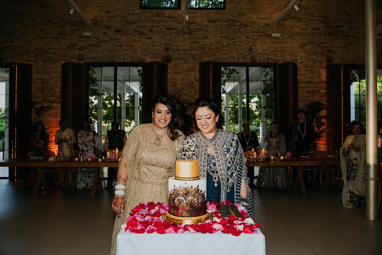 Two Indian brides playfully cutting their wedding cake together during the reception.