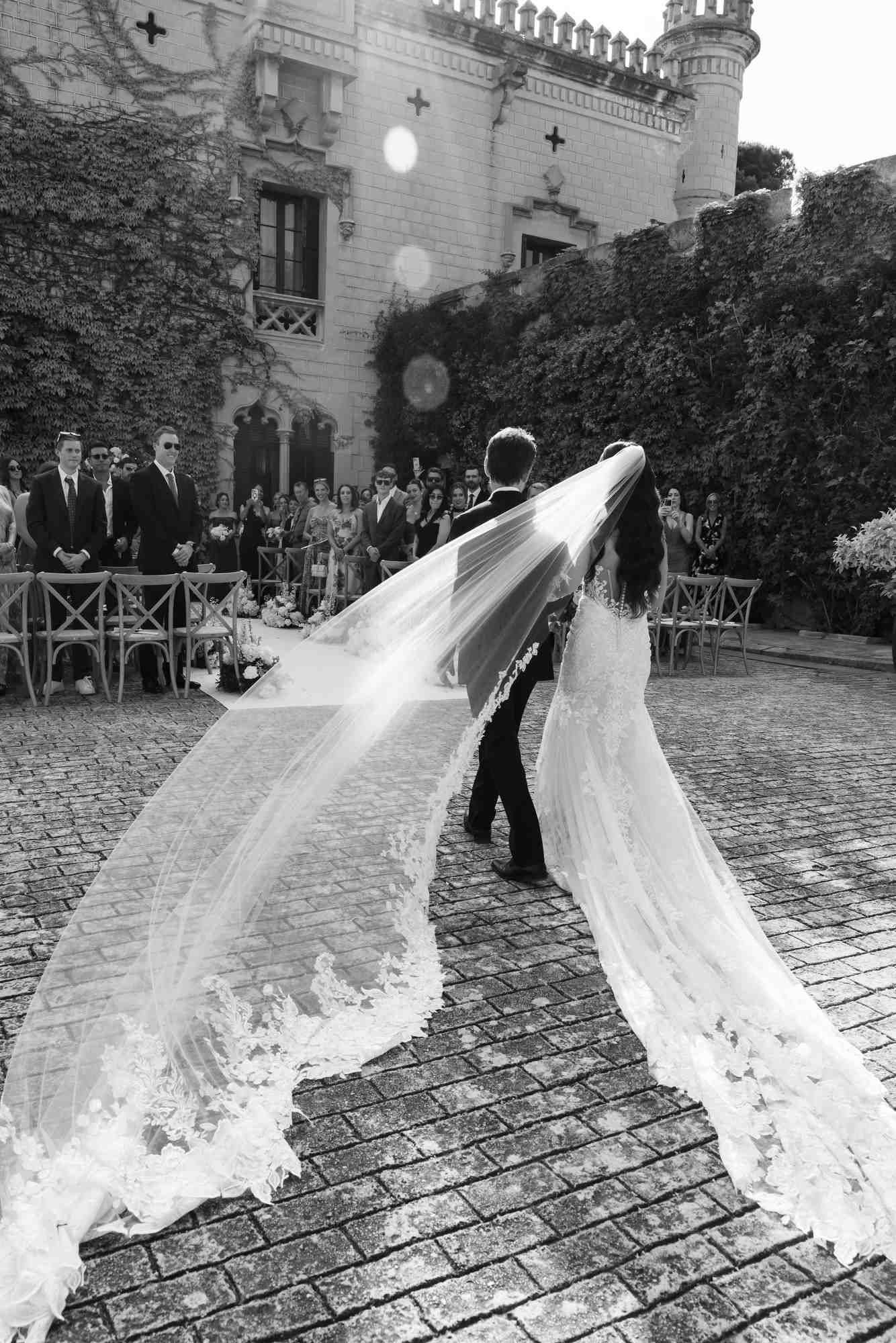 The bride and groom walking to their wedding ceremony at Castell Jalpí in Barcelona, as the wind catches the long veil